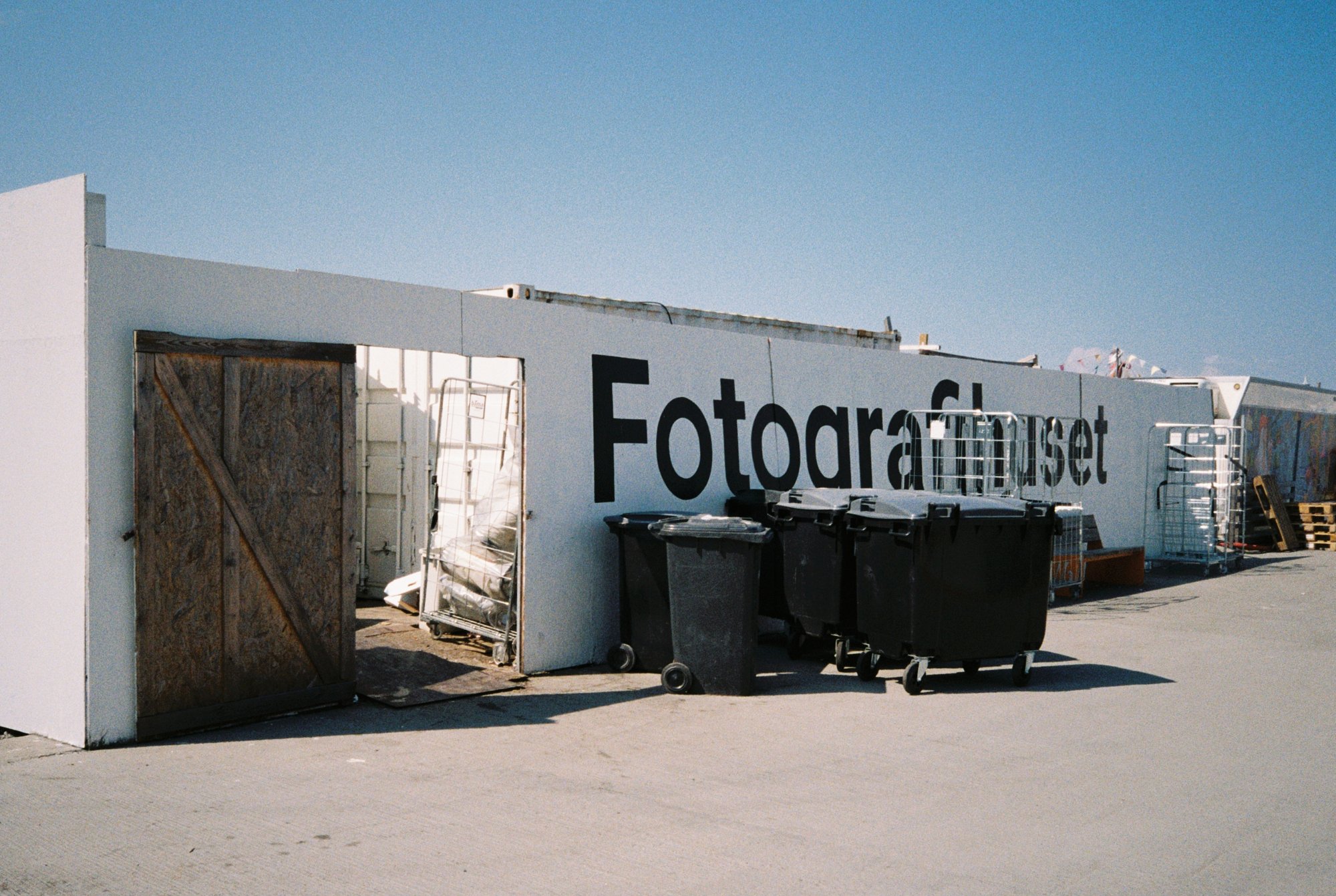 An outdoor scene with a white building wall that has black text reading 'Fotogra'. There are trash bins and carts near the wall, with a wooden door and metal racks, under a clear blue sky.