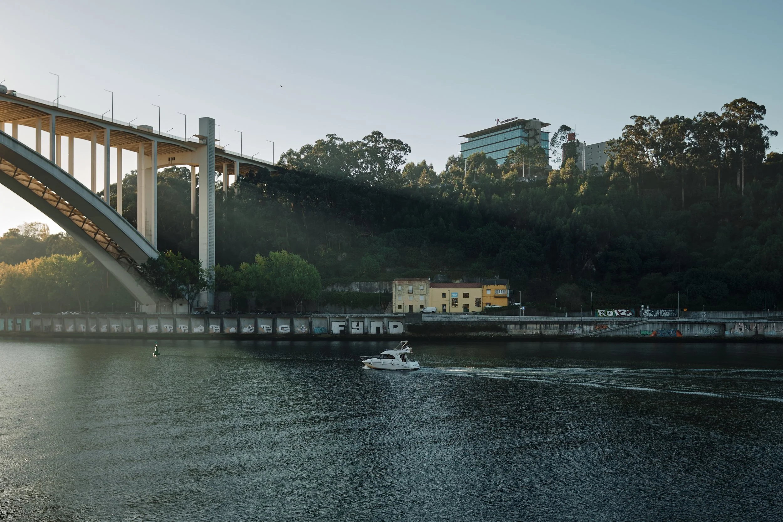 A boat sailing in a river with a hillside in the background, featuring a bridge, trees, and buildings.