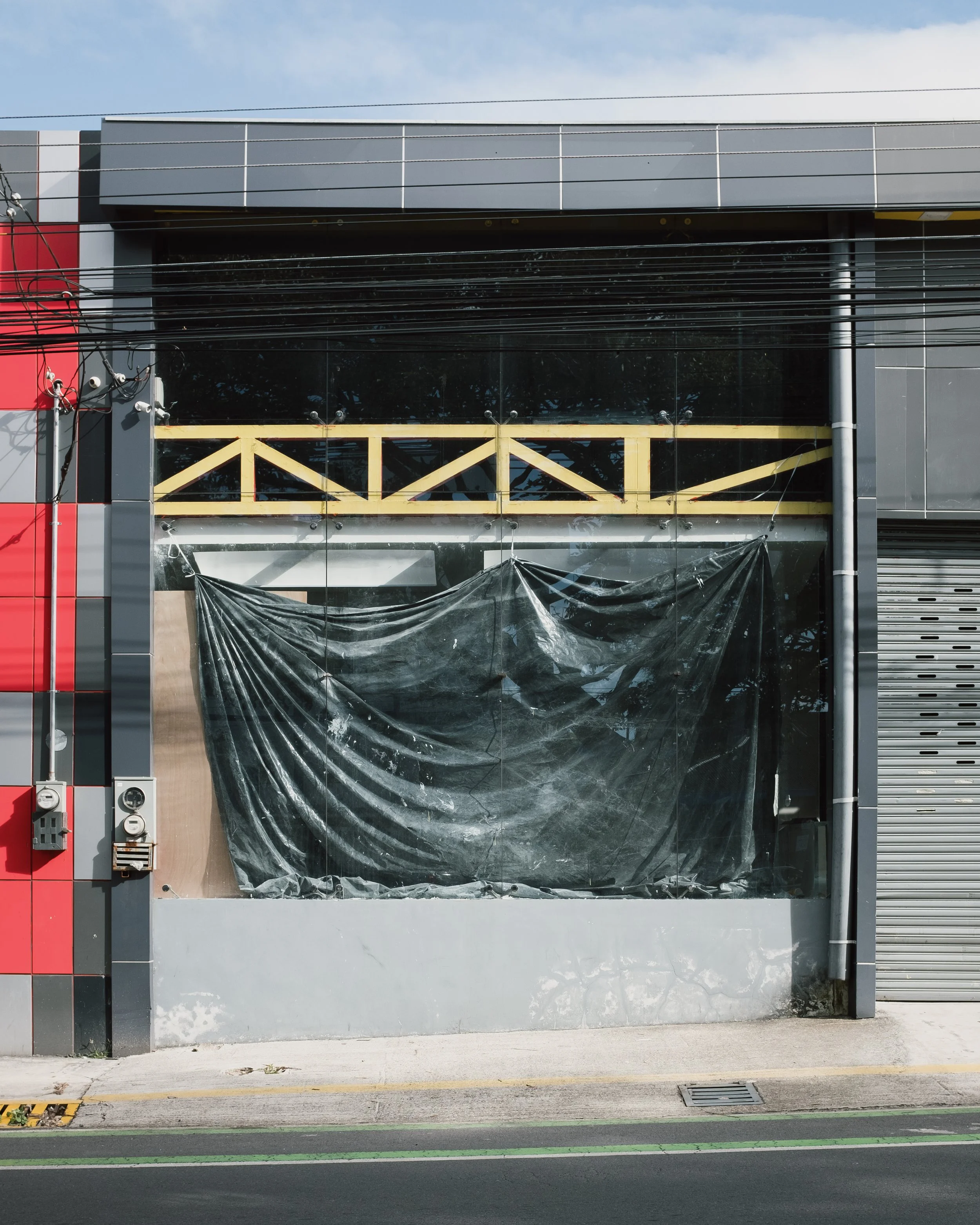 Empty storefront with large glass window covered by black plastic and yellow wooden beams inside, next to a gray roller shutter door.