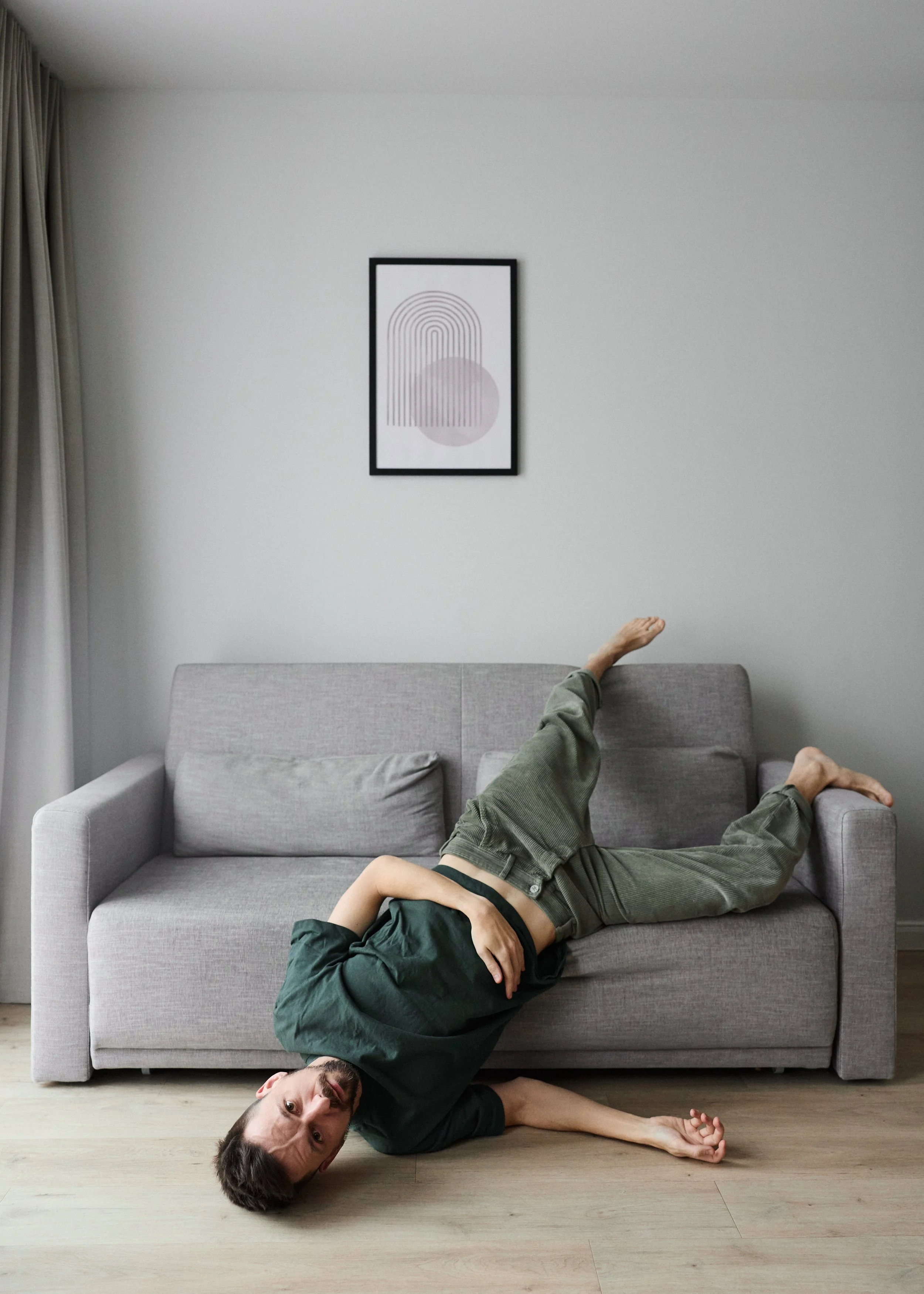 A man lying on a gray sofa with his head on the floor, one arm resting on his stomach, wearing a black t-shirt and green pants. One leg is draped over the back of the sofa while the other is bent at the knee. A picture with abstract lines and shapes hangs on a plain white wall above the sofa.