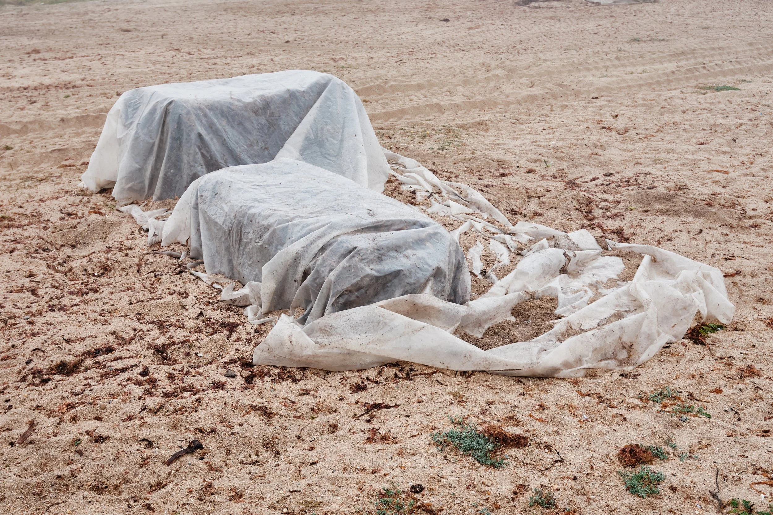 Objects covered with white and gray tarps lying on sandy ground in an open area.