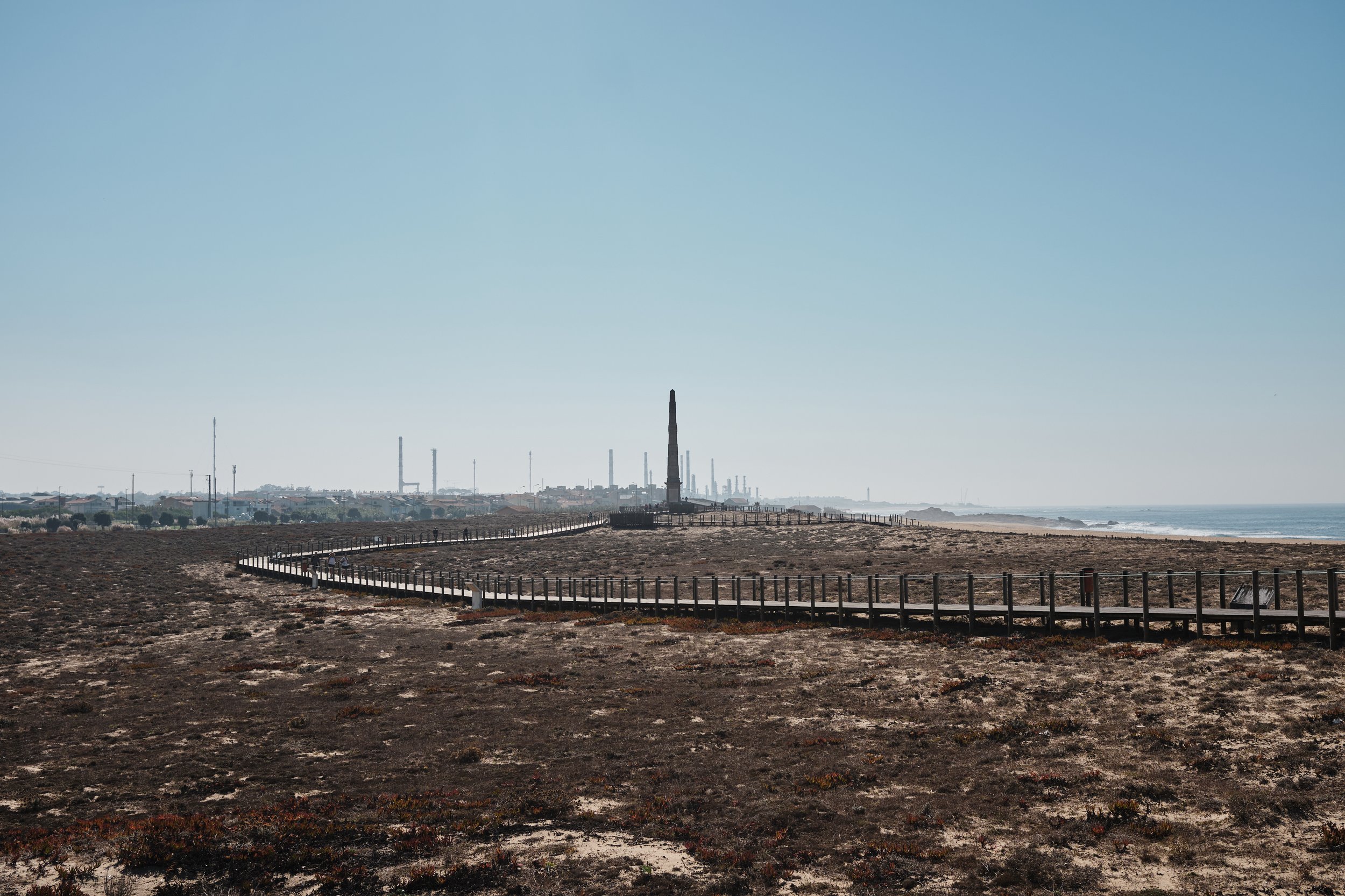 A coastal landscape with a wooden path leading towards a monument on the horizon, with industrial buildings and smokestacks in the background on a clear day.