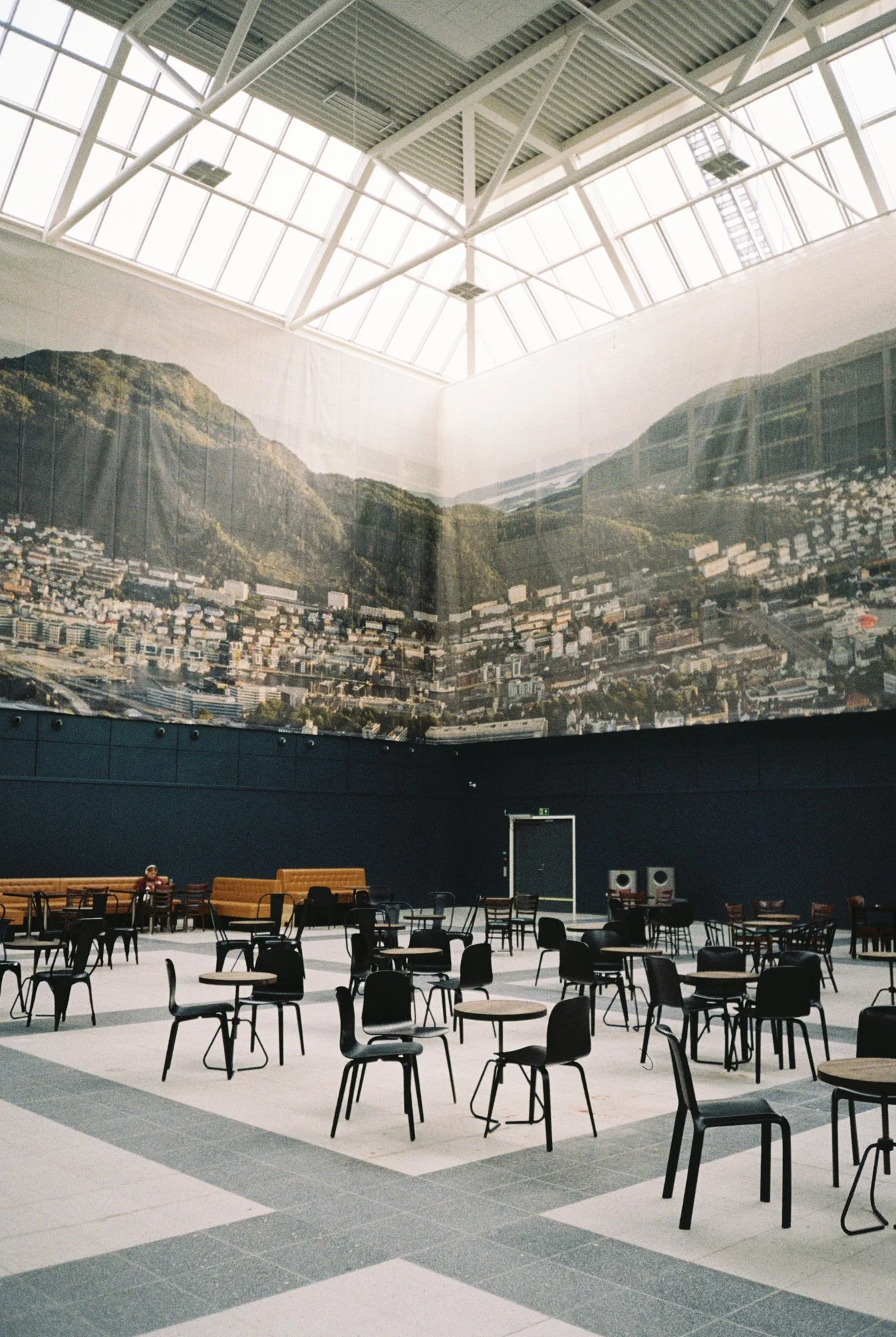 Empty indoor public space with black chairs and tables, large mountain mural on the wall, and a skylight ceiling.