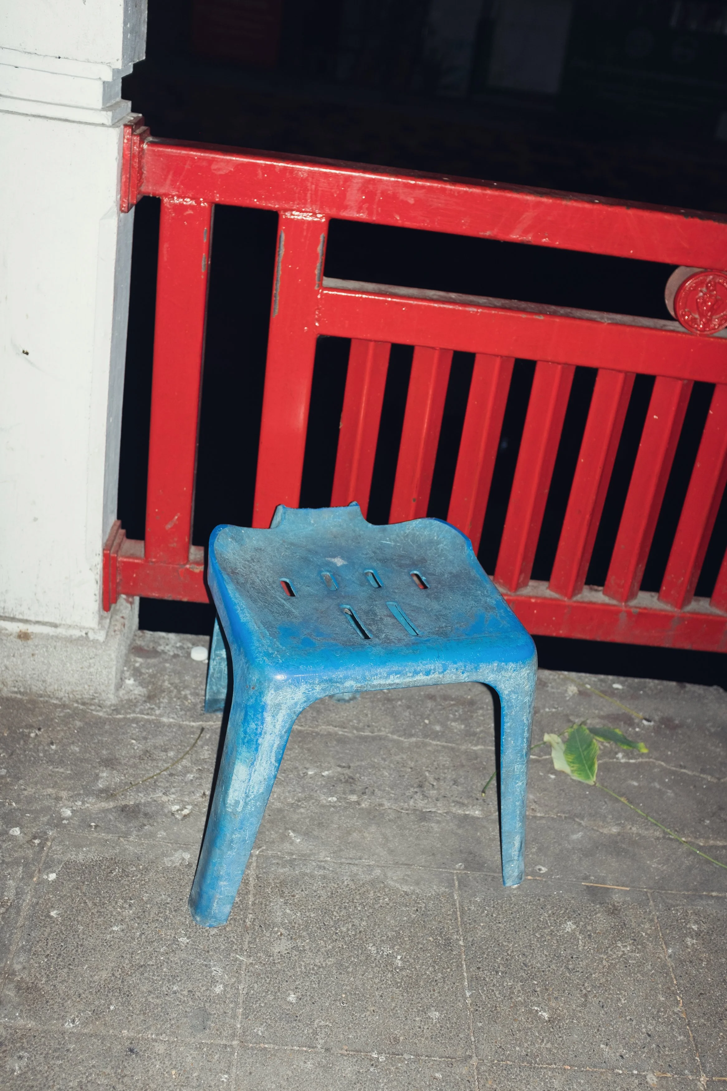 A small blue plastic stool situated on a concrete floor, with a red railing and a white column in the background.