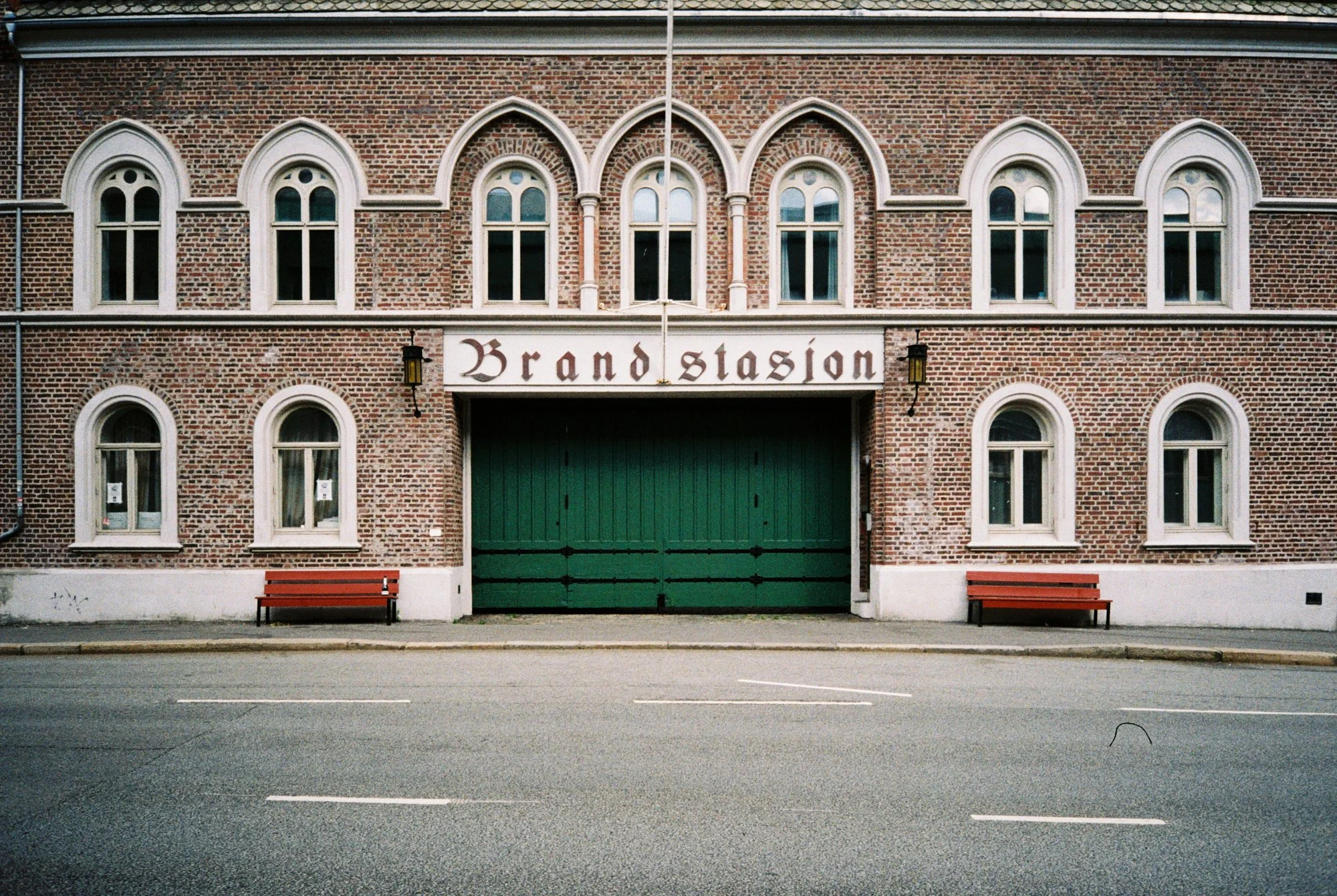 Brick building with arched white windows and a sign that reads 'Brand Stasjon' above a large green gate. There are two red benches in front of the building and a road with lanes in the foreground.