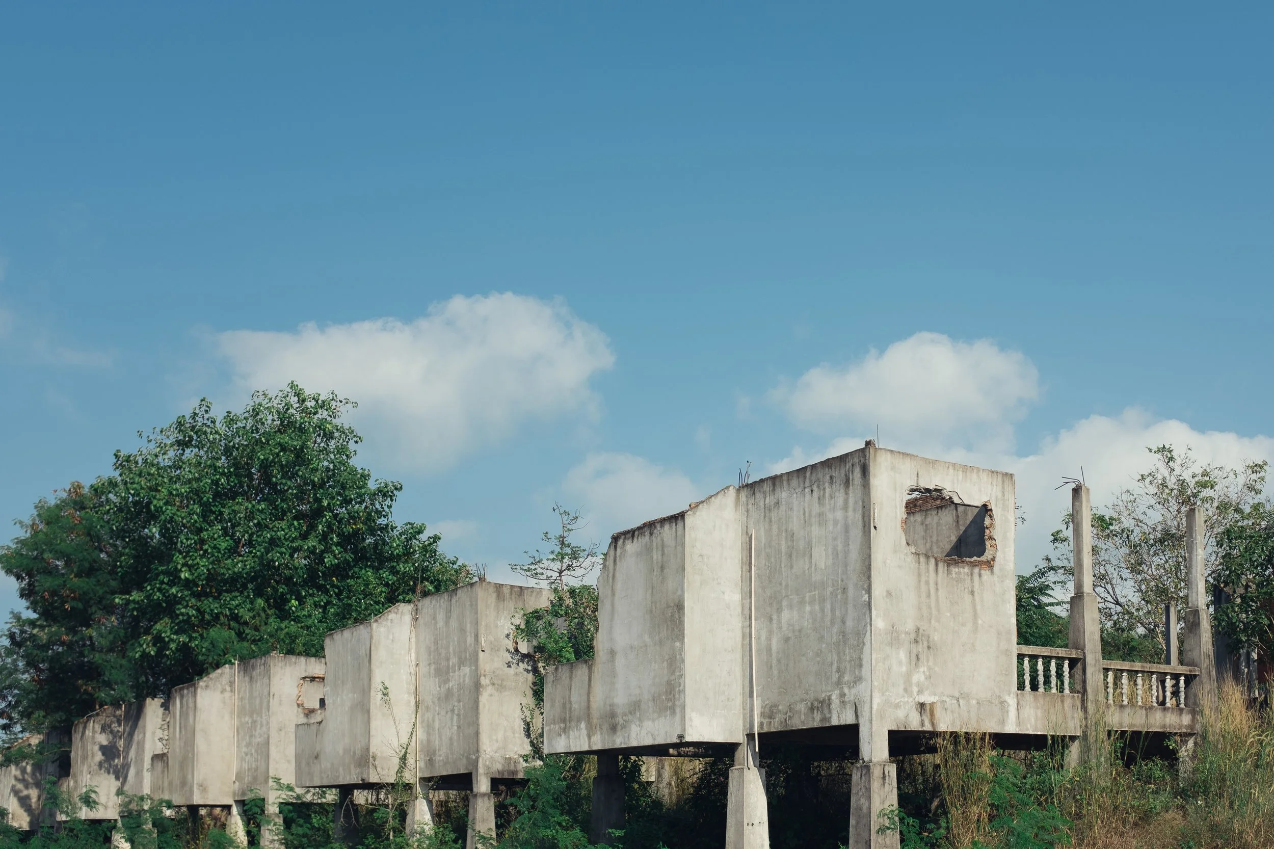 Abandoned concrete building with missing roof and broken windows, surrounded by green trees and under a blue sky with white clouds.
