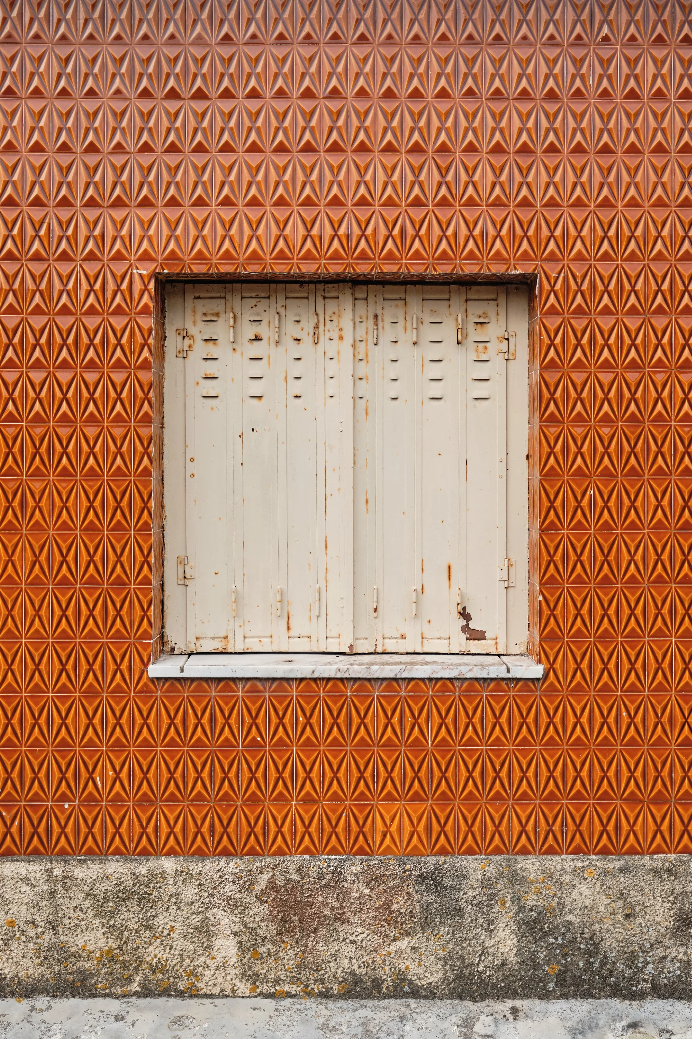 A window with white, weathered shutters set in an orange, geometric, textured tiled wall and a concrete base.