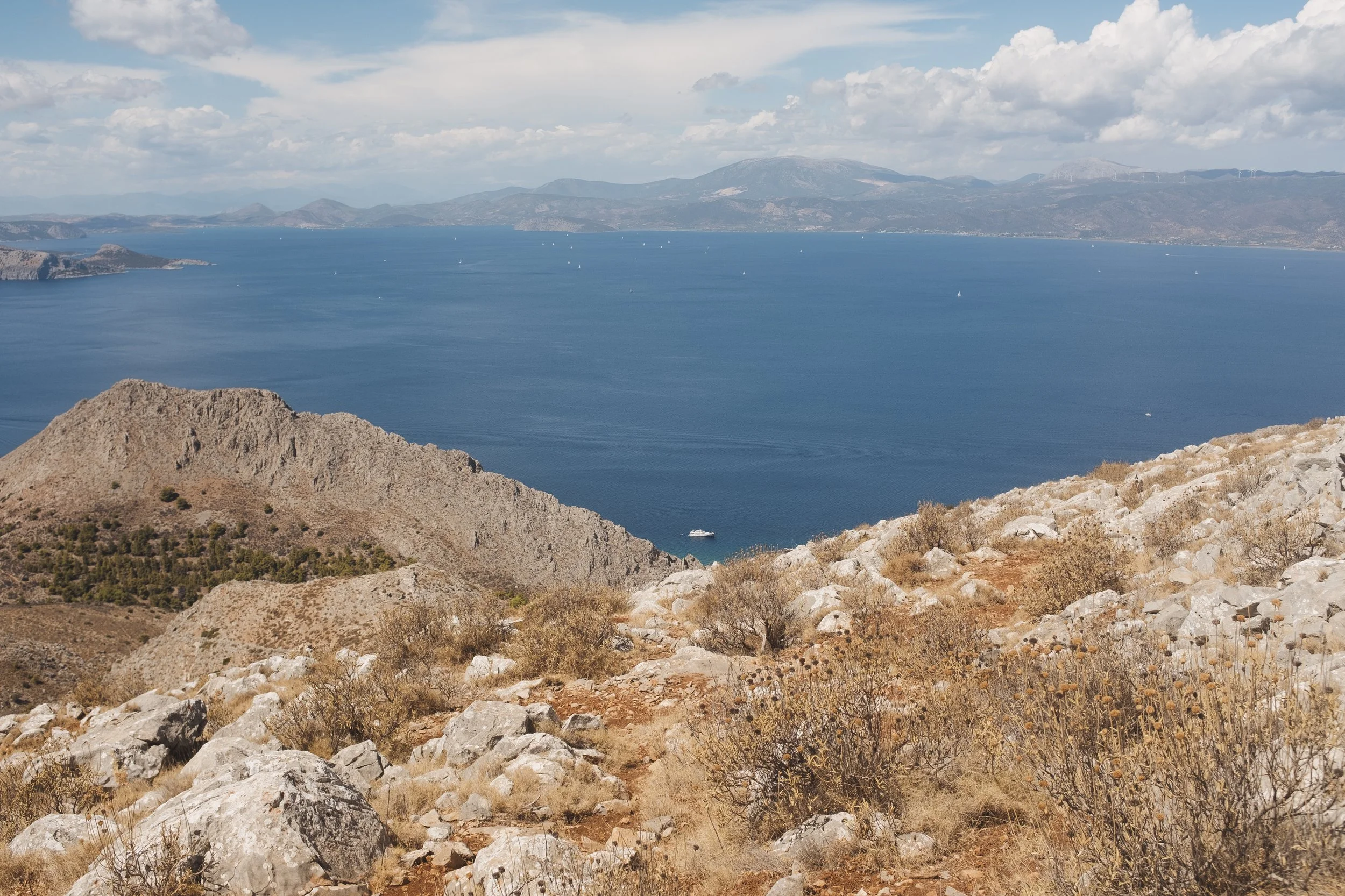 A scenic view of a large blue lake from a rocky hillside with sparse shrubs, surrounded by mountains with partly cloudy skies.