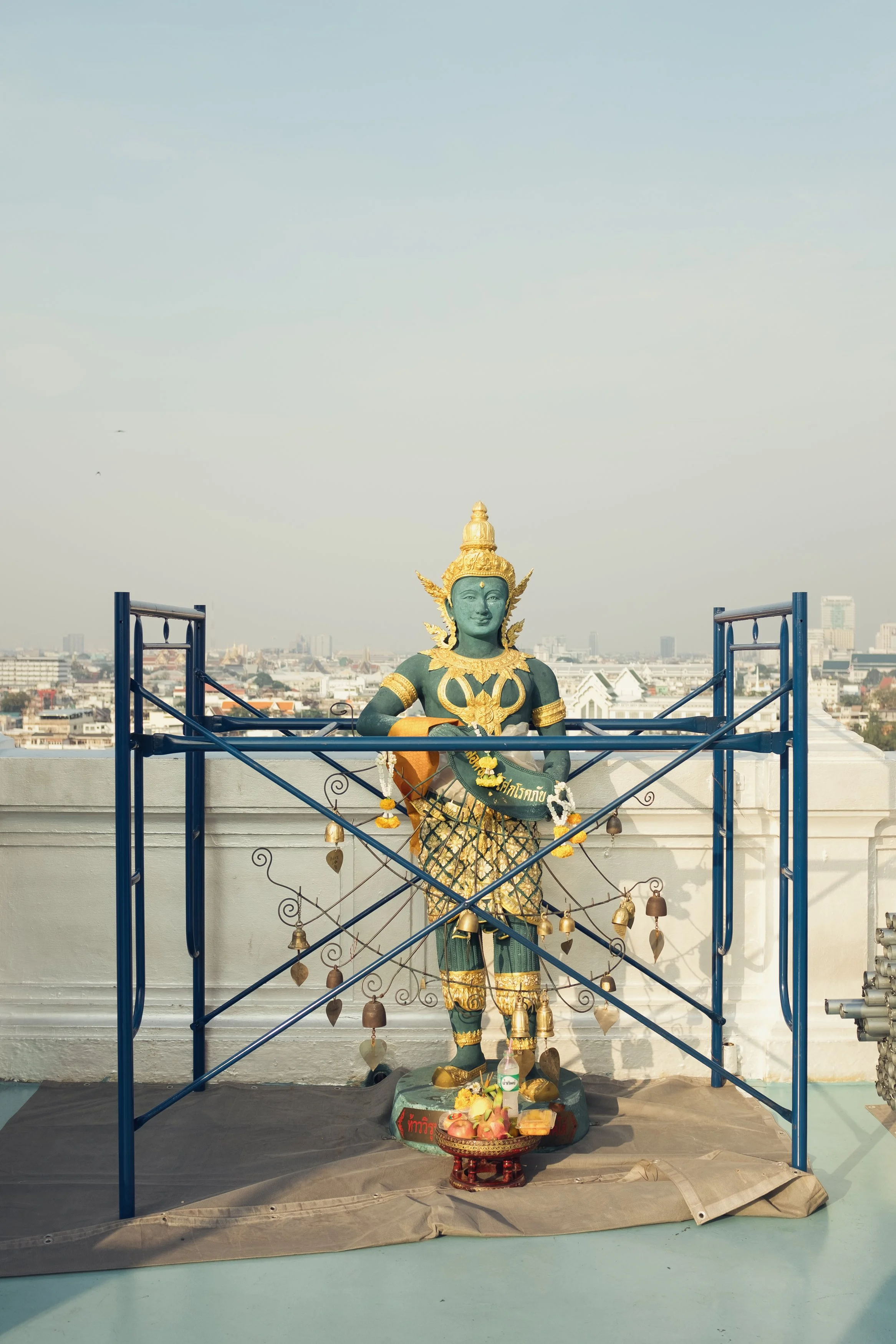 Statue of a Hindu deity surrounded by a metal frame with bells, set on a rooftop against a cityscape background.