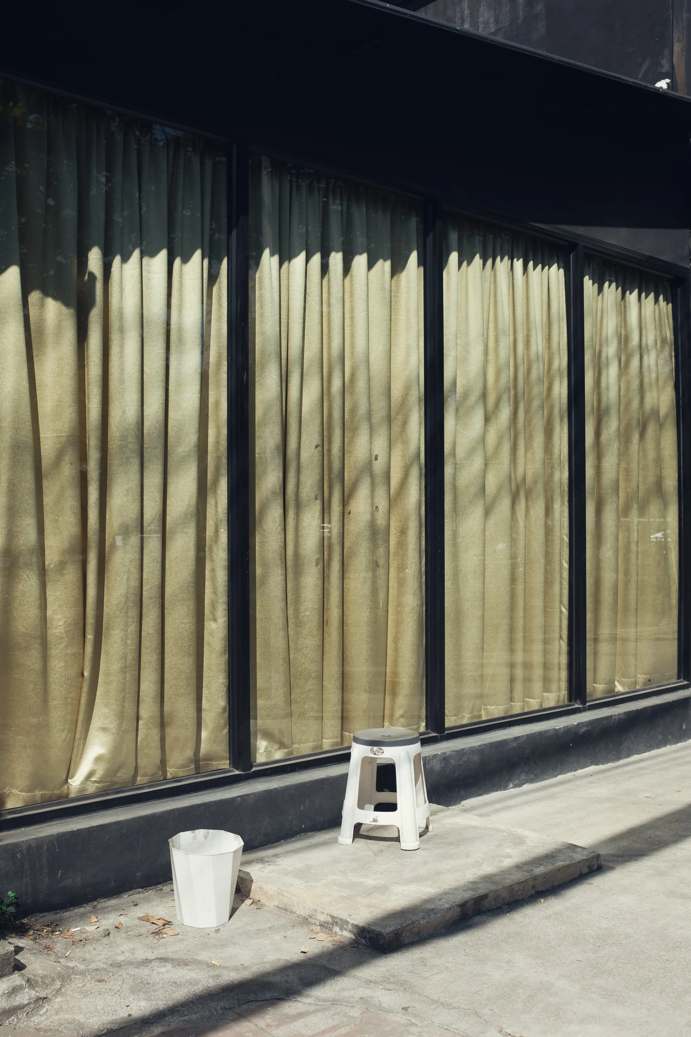 A large window with beige curtains and black frame, with a small white plastic stool and bucket outside on the sidewalk.