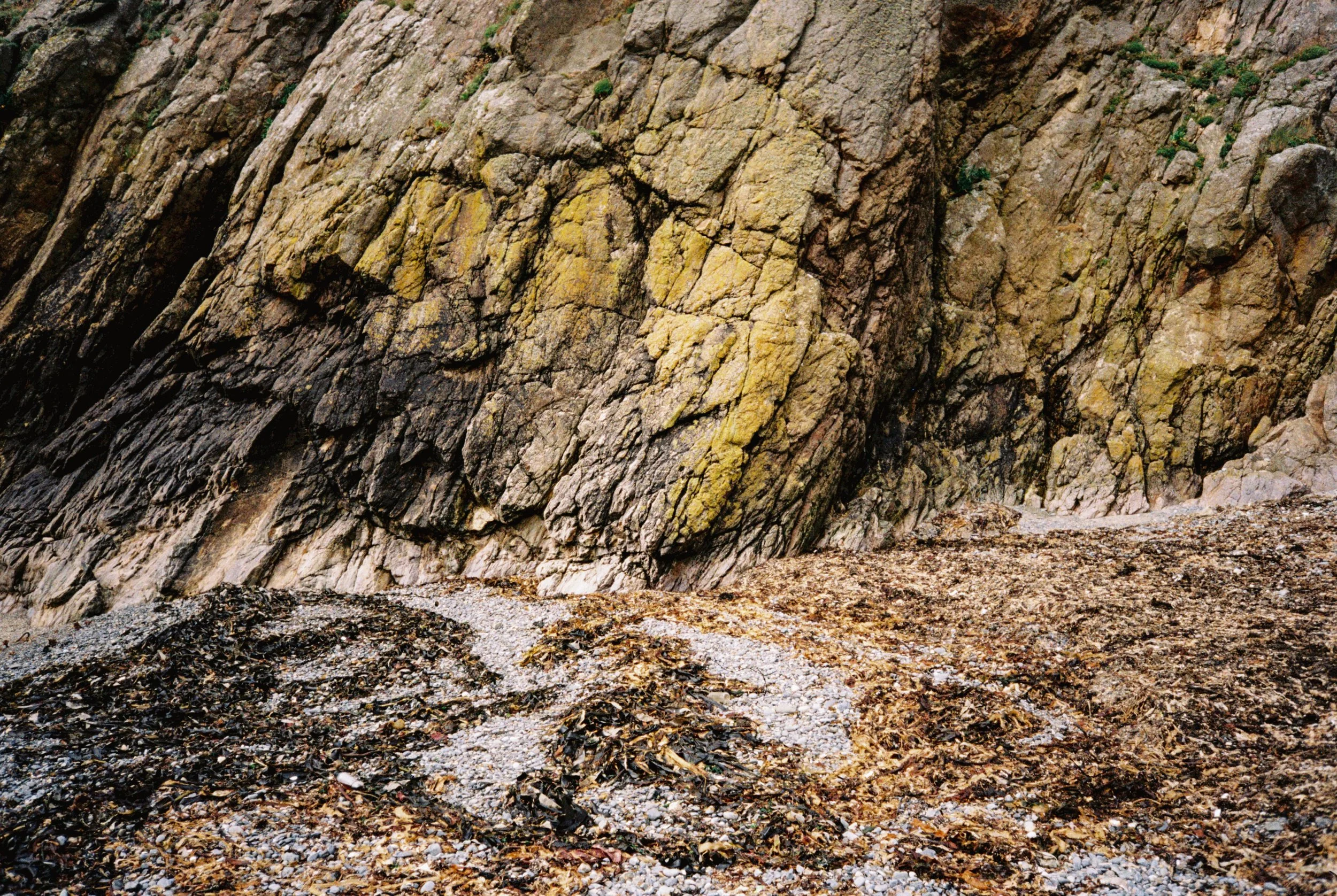 Close-up view of a rocky cliff with a mixture of black, yellow, and gray rocks and some seaweed on the ground at the base.