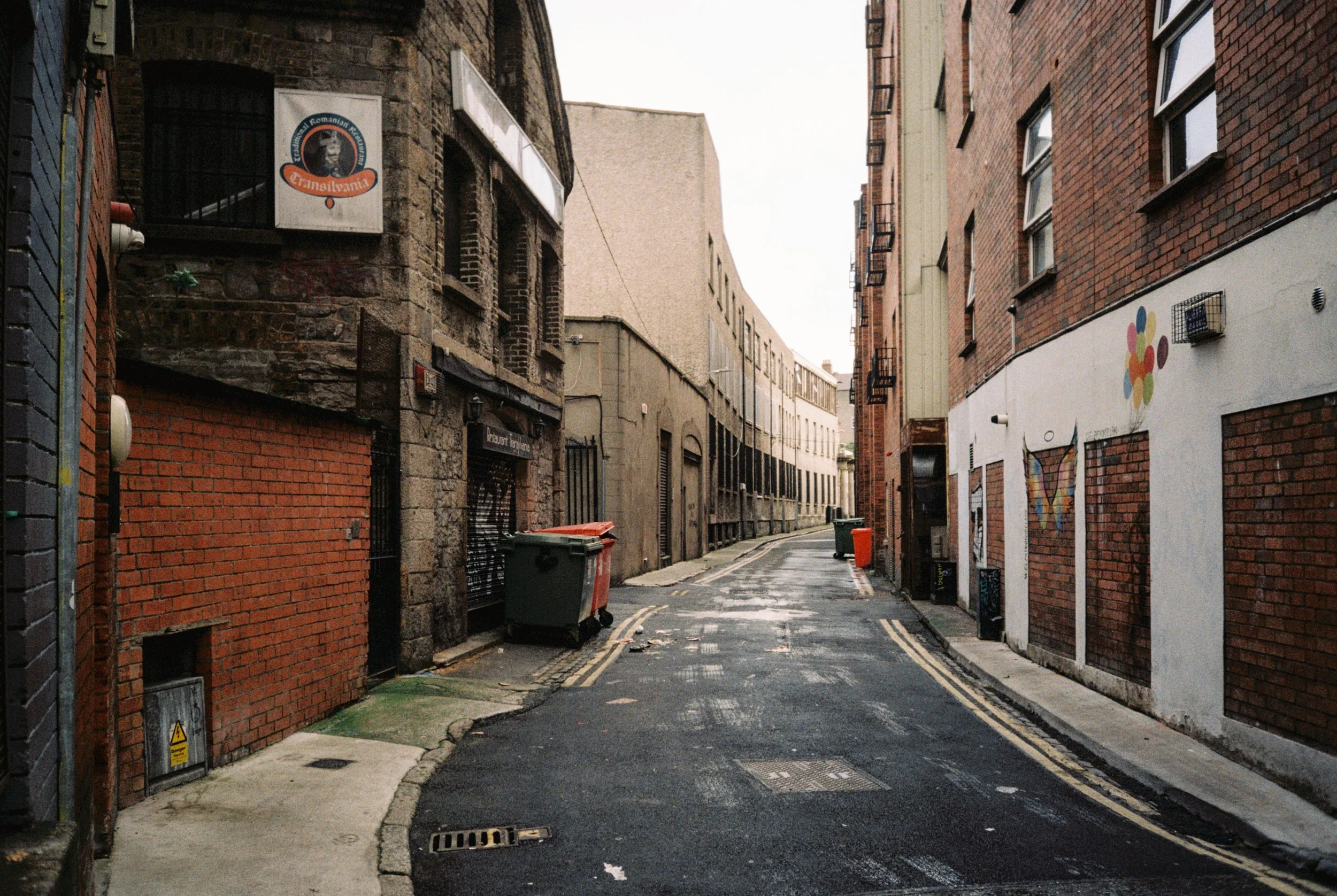 Empty narrow city street with brick and stone buildings on both sides, some with graffiti art, trash bins, and a wet road, under an overcast sky.