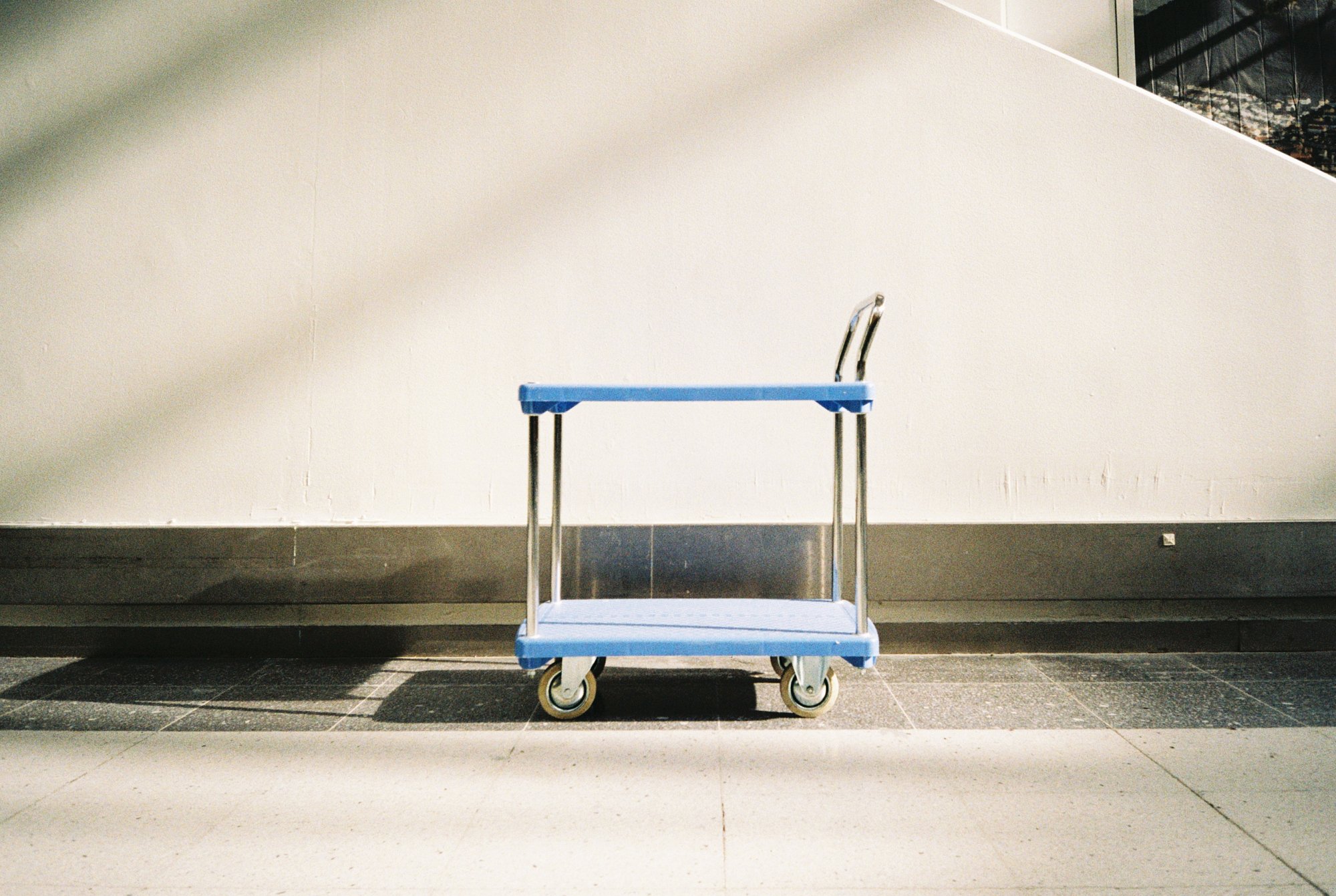 A blue utility cart with a handle and four wheels, empty, positioned against a beige wall on a tiled floor.