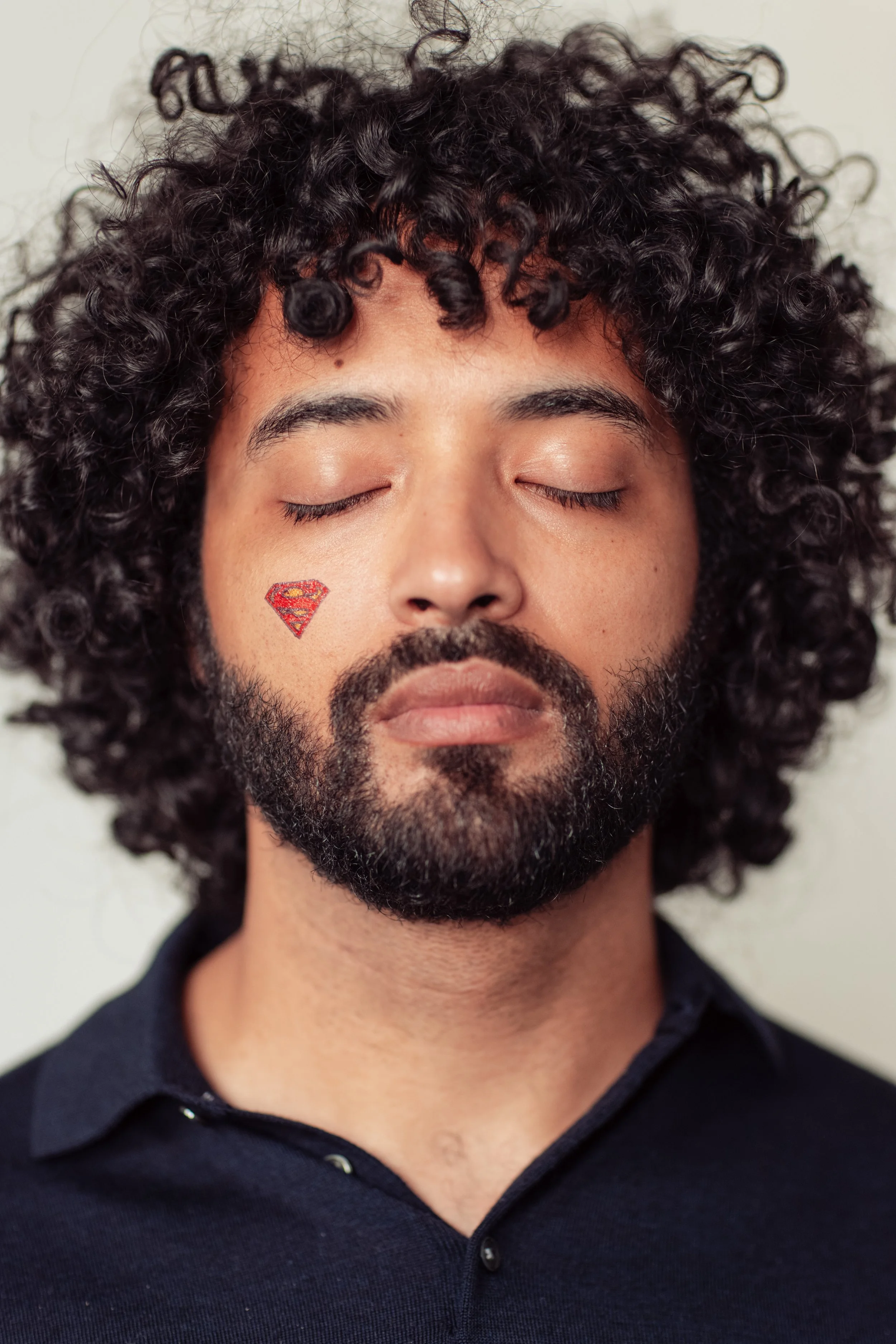 A man with curly hair and a beard has his eyes closed. There is a Superman logo sticker on his cheek.