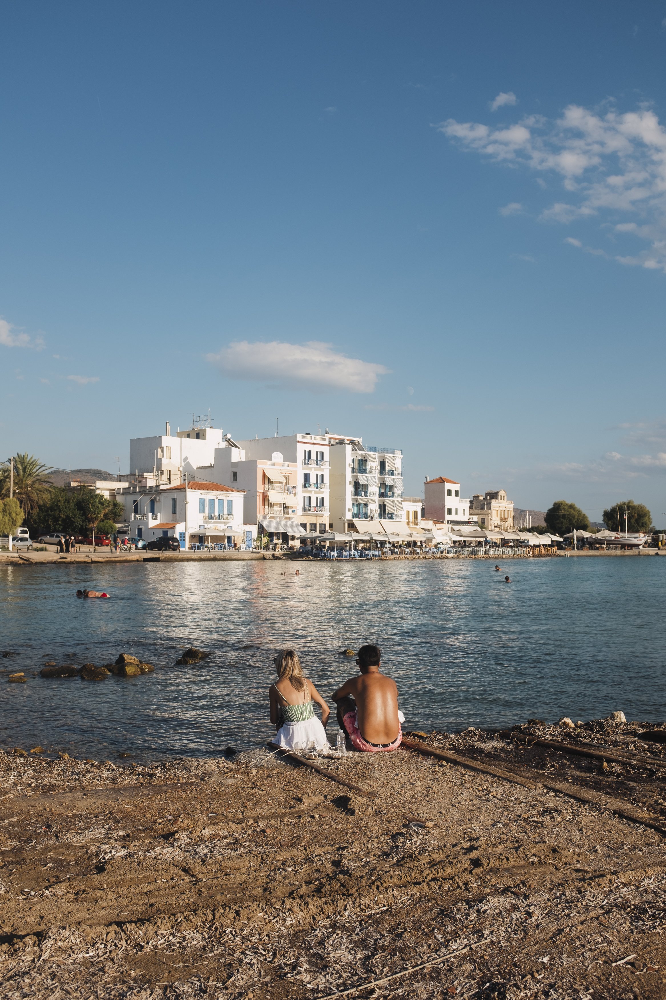 Two people sitting on a rocky shore looking at the water with white buildings and blue sky in the background.