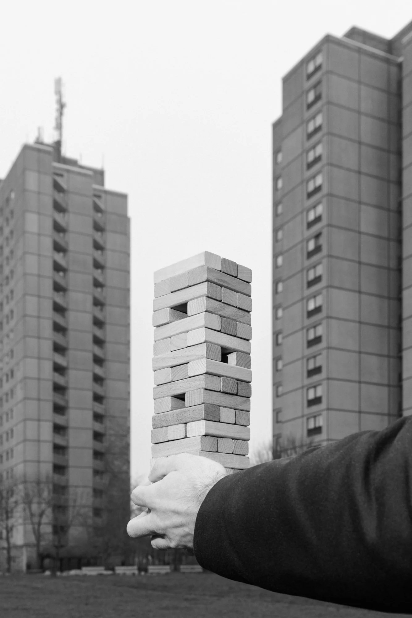 A person holding a large Jenga game tower outdoors, with tall apartment buildings in the background.