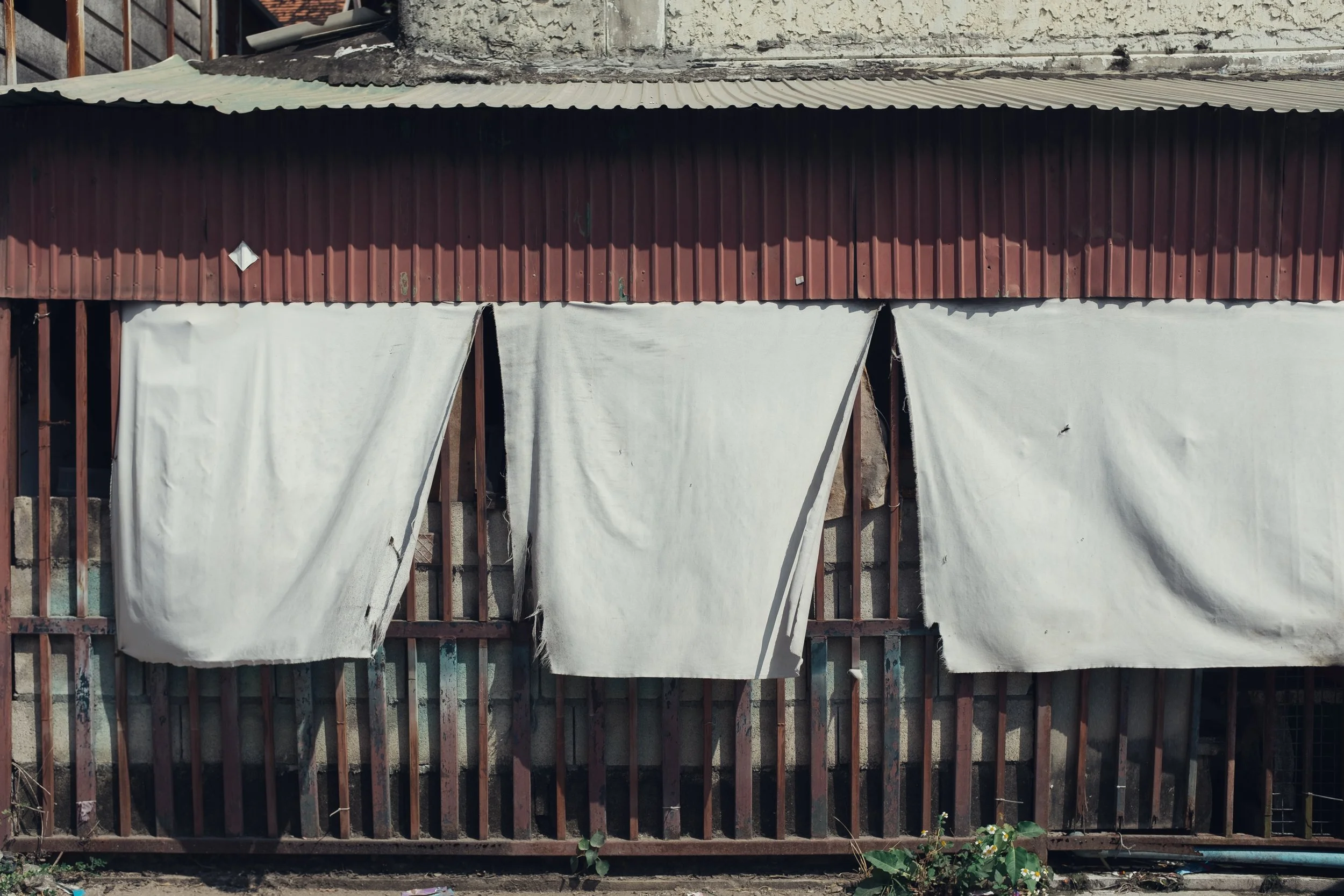 An old building with a metal roof, red corrugated siding, and white fabric curtains hanging in front, partially covering the structure.