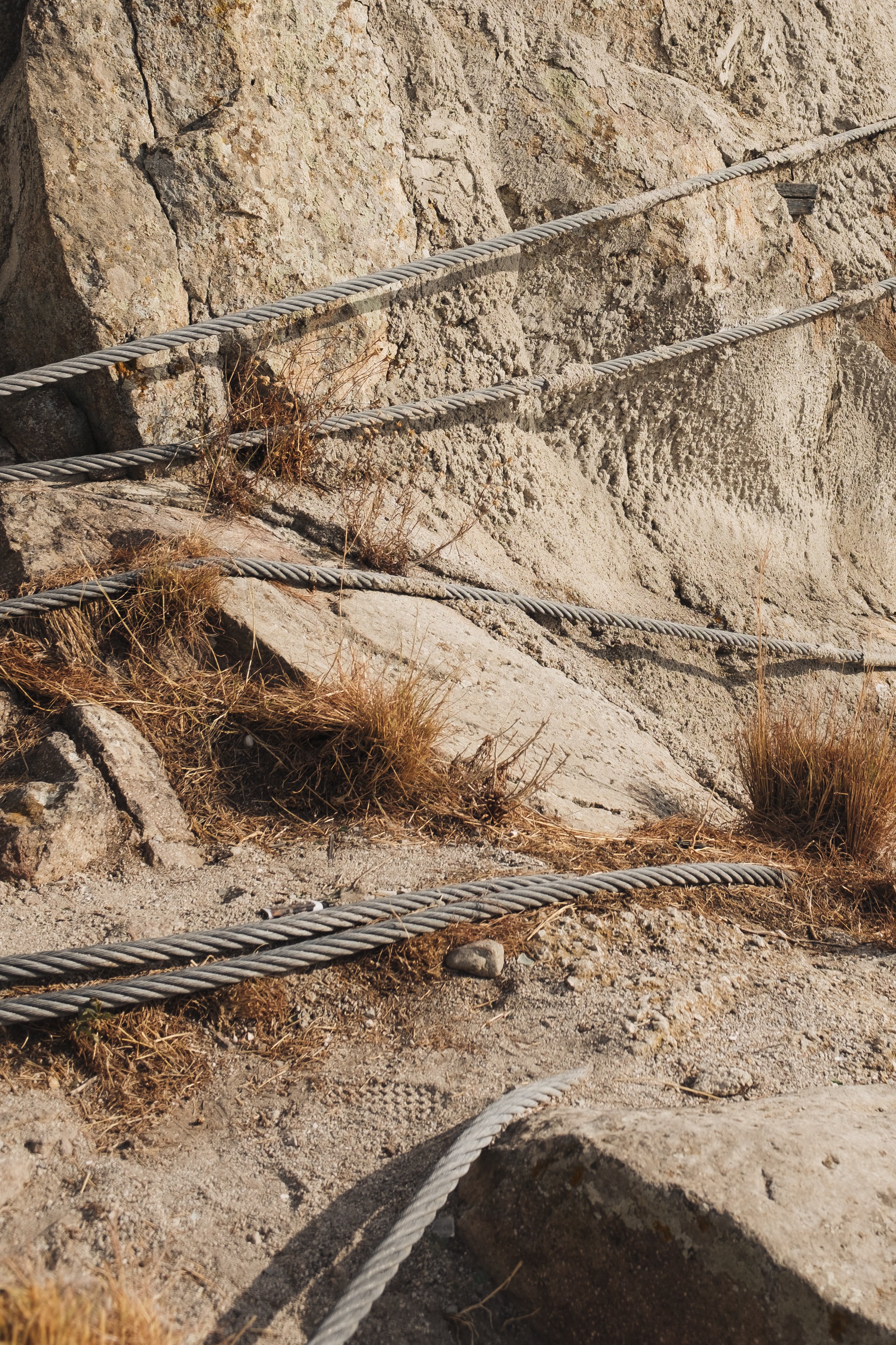 Close-up of rugged mountain trail with metal cable handrails and dry grass, rocky terrain, and sunlight shadows.