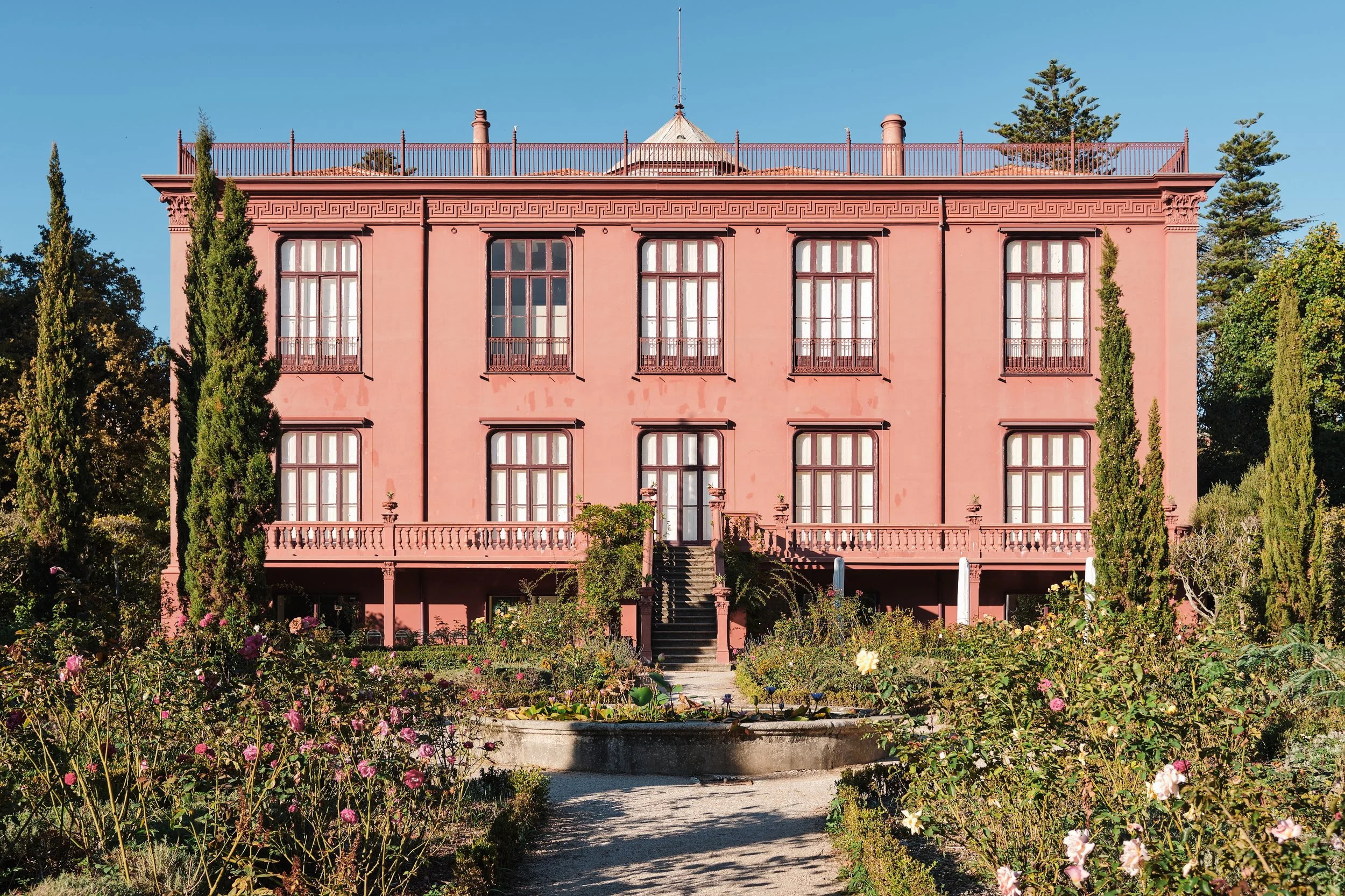 A three-story pink mansion with large windows, a balcony on the second floor, and a staircase leading to the front door, surrounded by a garden with rose bushes and tall trees under a clear blue sky.