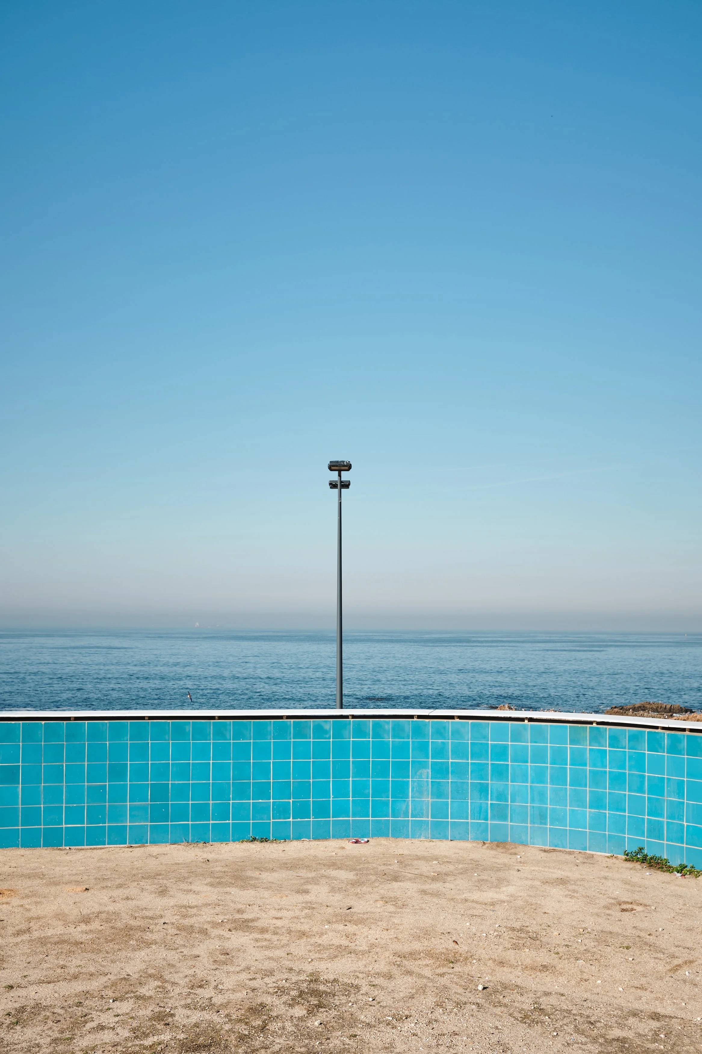 View of a coastal scene with a blue sky, ocean, a tall lamppost, and a sandy area with a blue tiled wall in the foreground.
