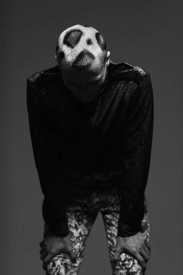 Person with hair dyed white and black spots, bowing with hands on knees, wearing a shiny black jacket and patterned pants, in a dark studio setting.
