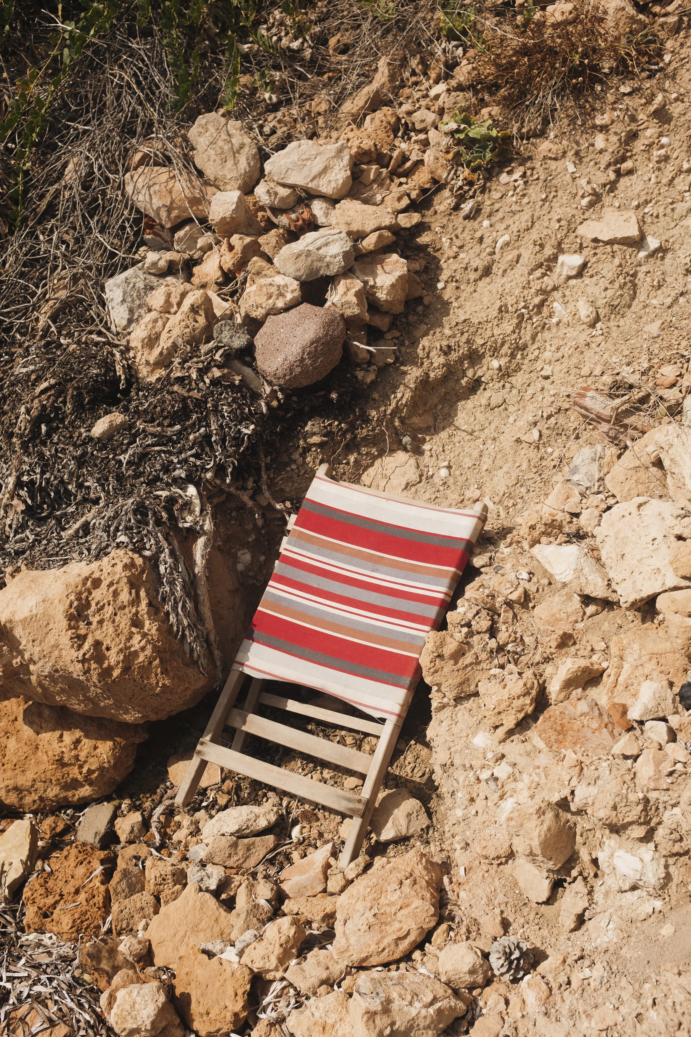 A small wooden deck chair with a striped fabric seat in red, gray, and white, resting on rocky, sandy terrain in an outdoor, desert-like environment.