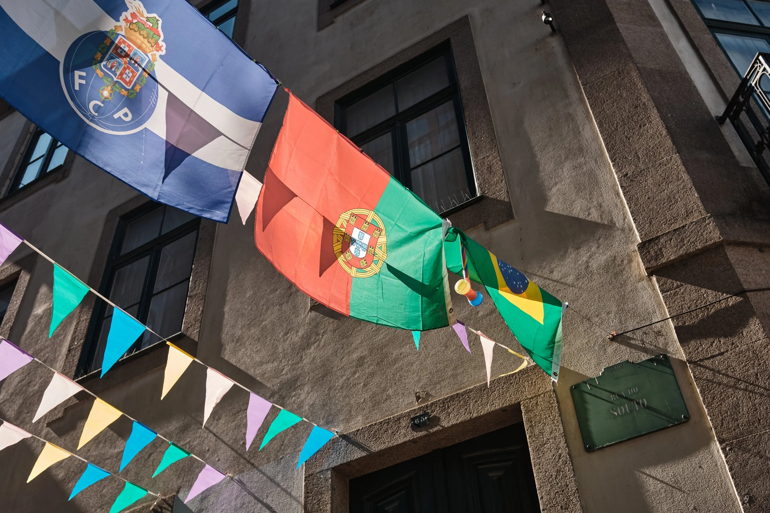 Flags of Portugal, Brazil, and other colorful flags hanging above the street in front of a stone building.