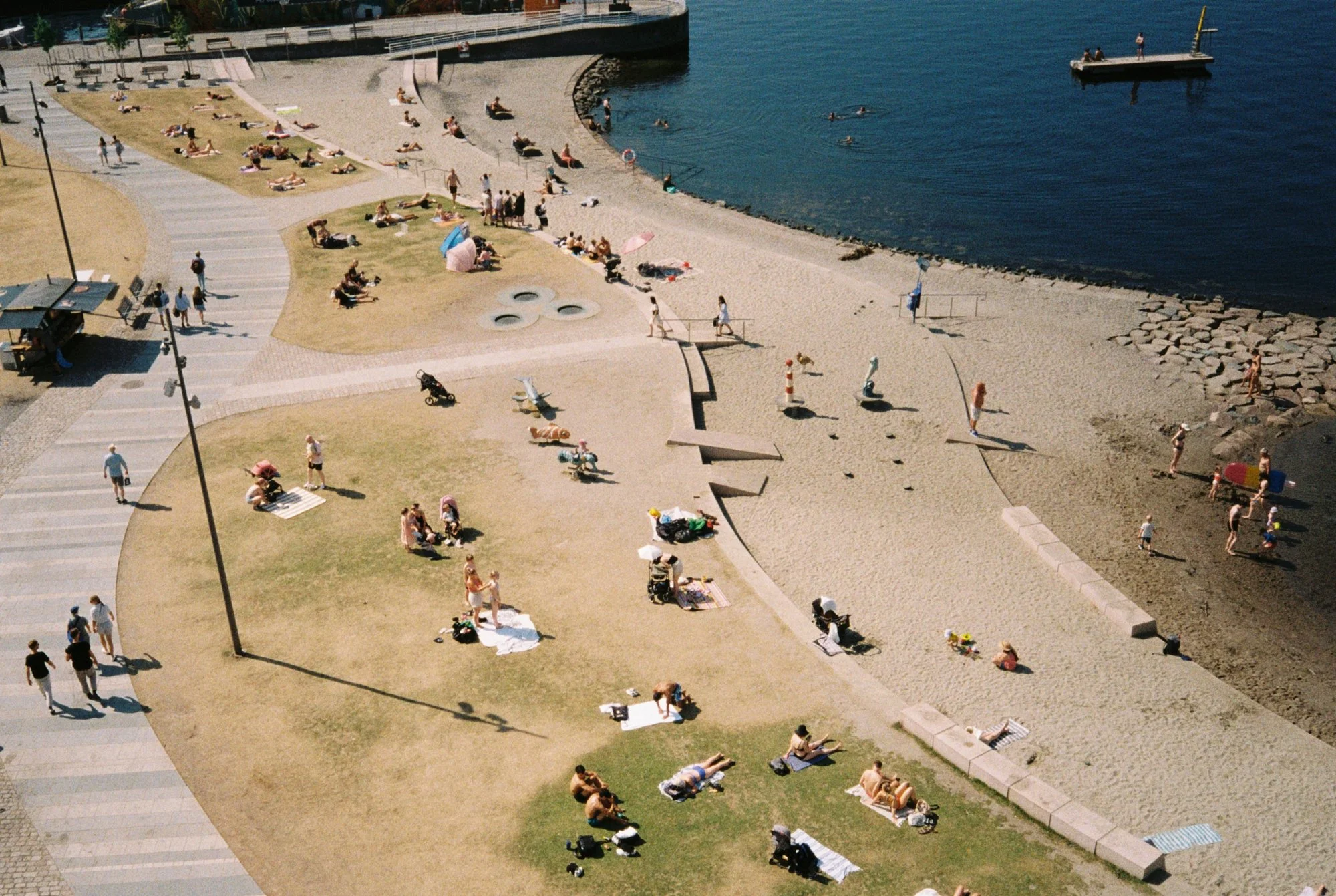 People sunbathing and swimming at a lakeside beach with grassy areas, a sandy shoreline, and a dock extending into the water.