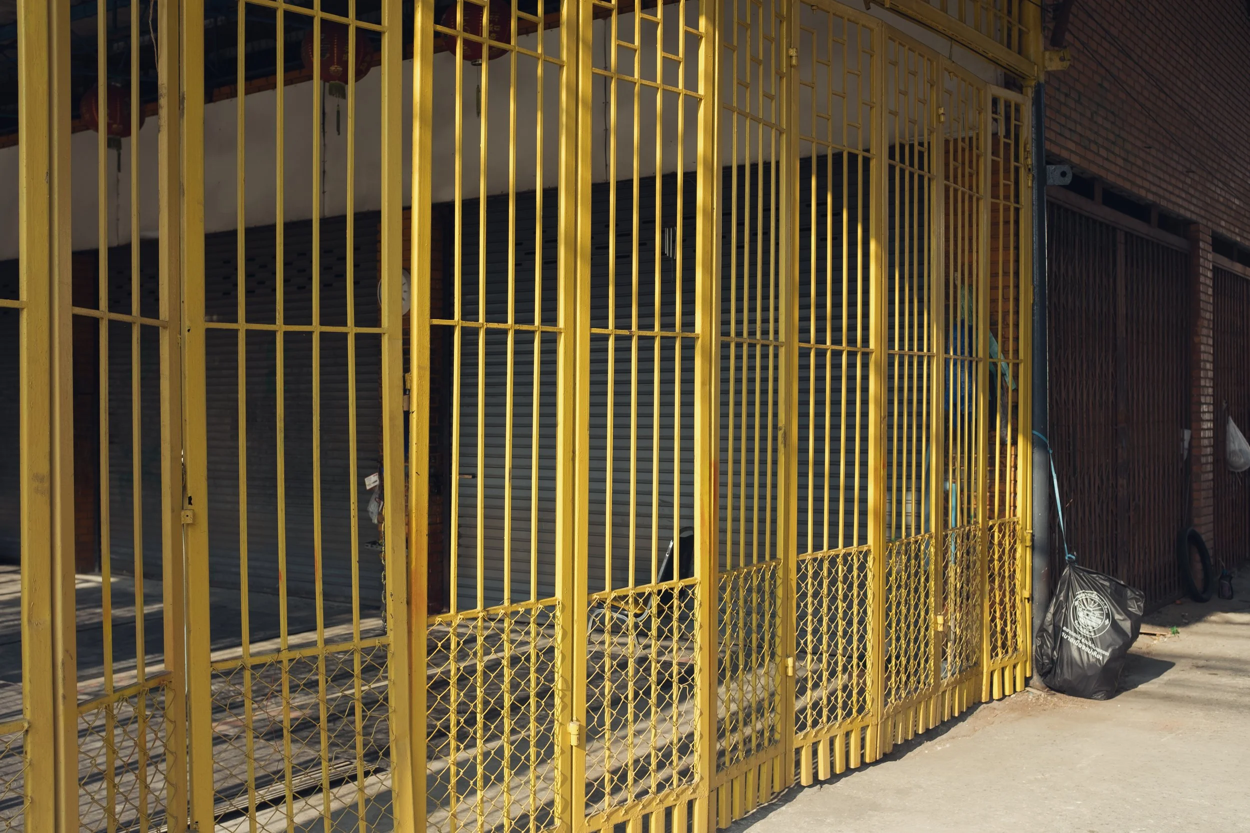 Closed yellow metal security gate in front of a storefront with a brick wall on the right side and a black plastic bag on the sidewalk.