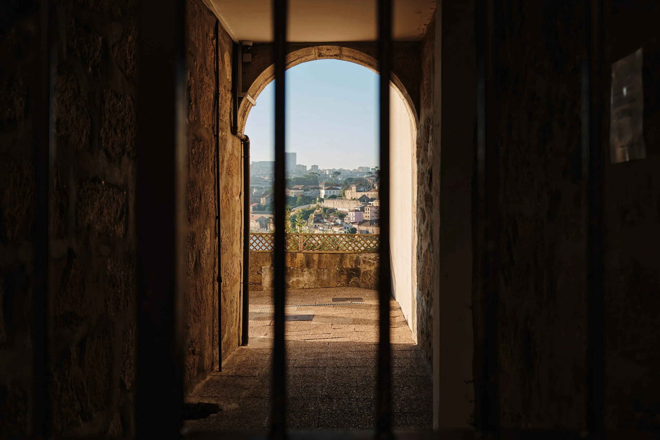 View of a cityscape through an arched opening seen from behind metal bars, with a stone wall and a railing in the foreground.