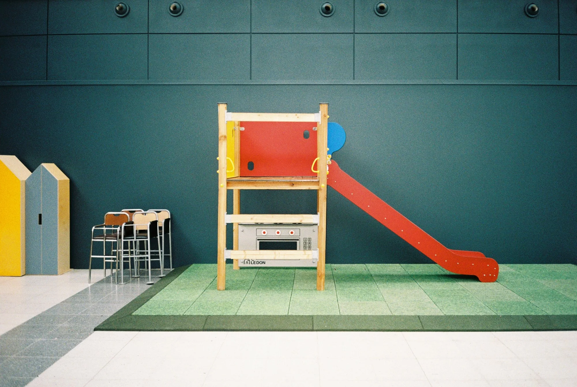 Empty indoor children's play area with a small red slide attached to a wooden structure, green rubber mat flooring, and stacked chairs to the left.