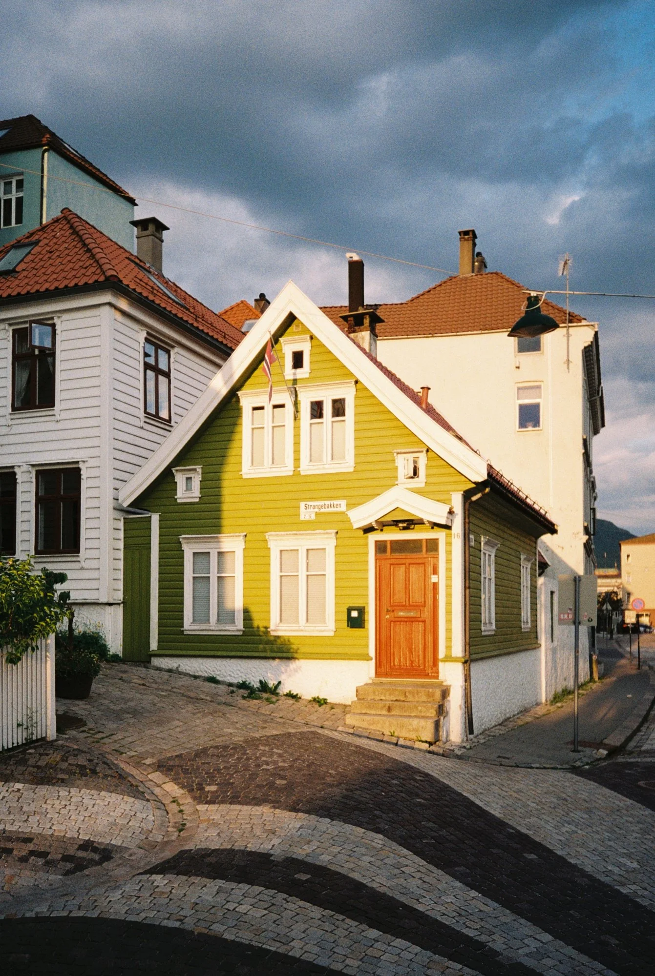 A green wooden house with white trim, a brown door, and multiple windows, situated on a cobblestone street with other houses nearby under a cloudy sky.