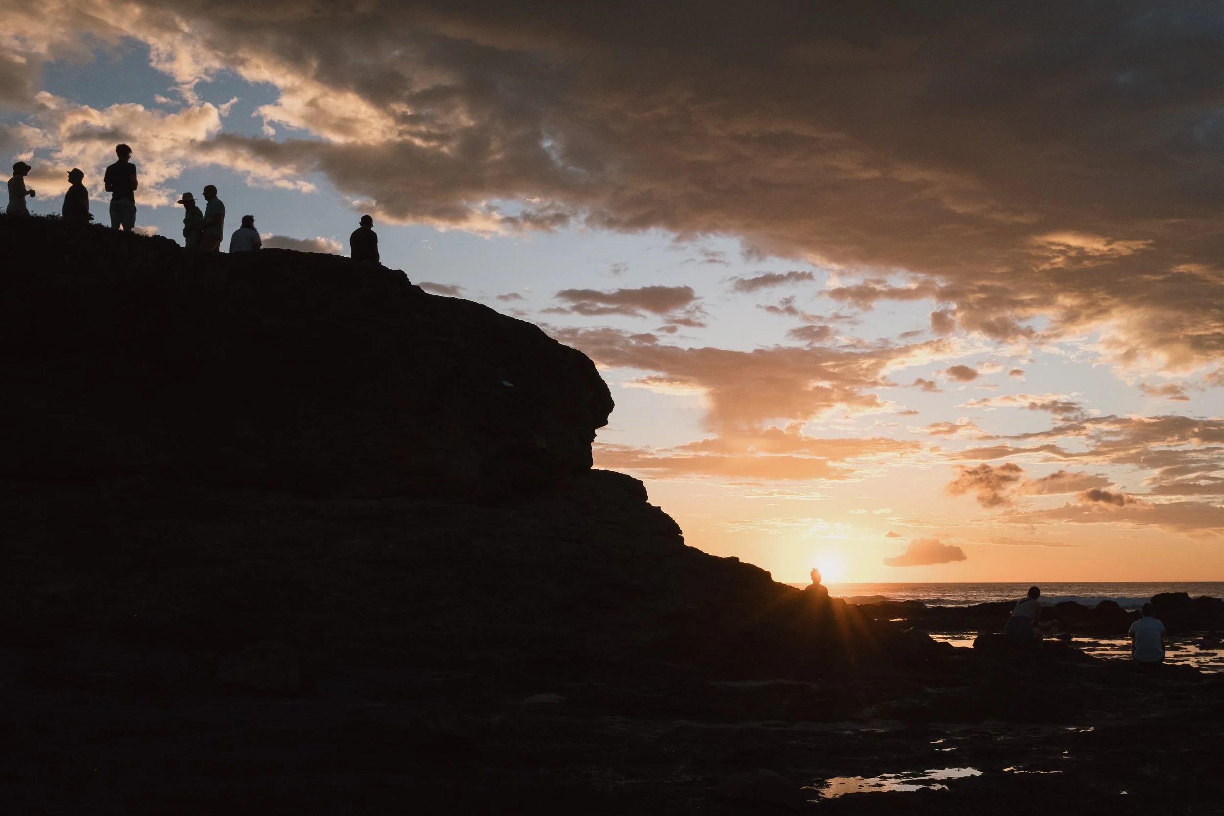 Silhouettes of people sitting and standing on a rocky cliff observing the sunset over the ocean, with clouds in the sky.