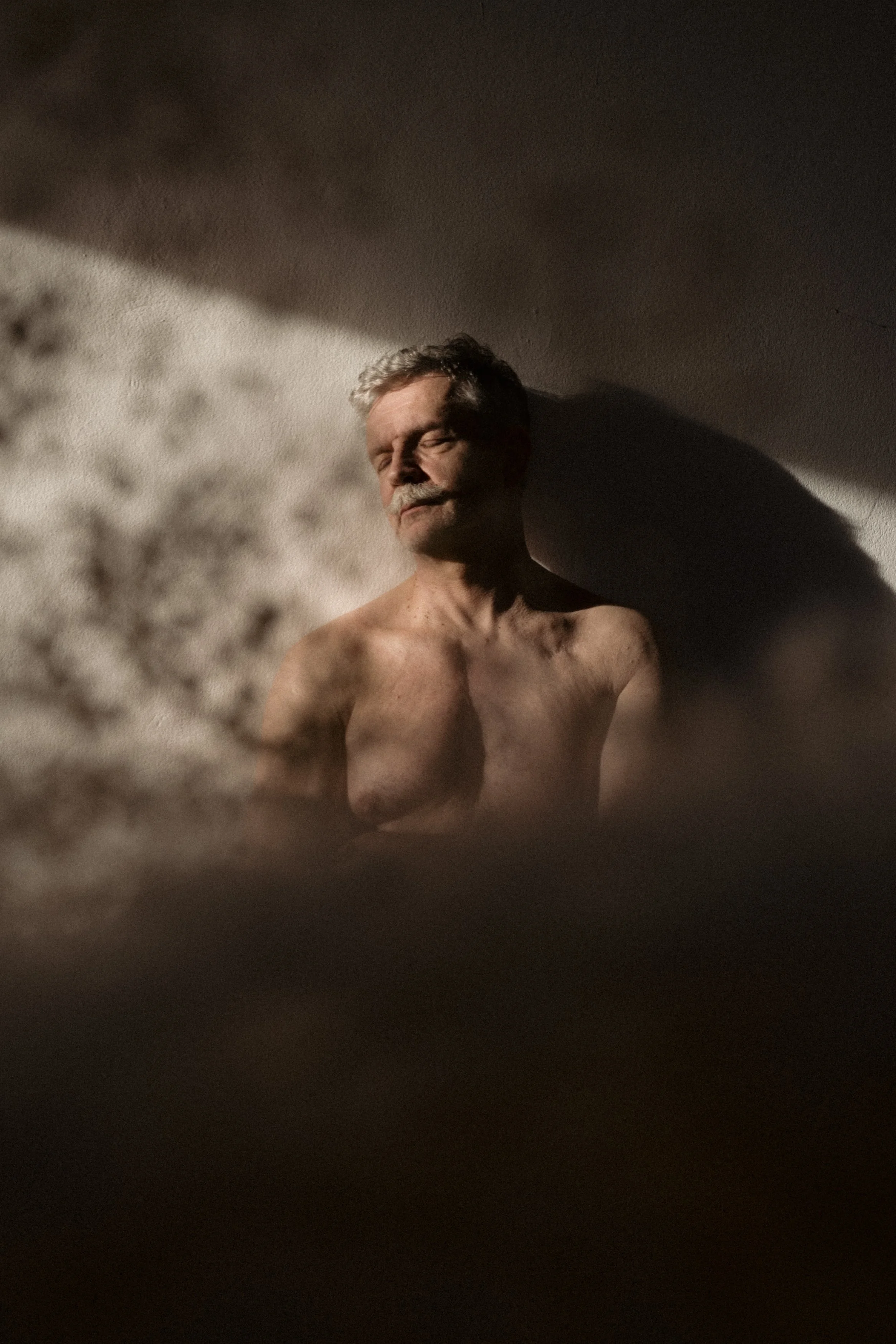 A shirtless elderly man with closed eyes, standing against a textured wall with soft lighting casting shadows on his face and body.