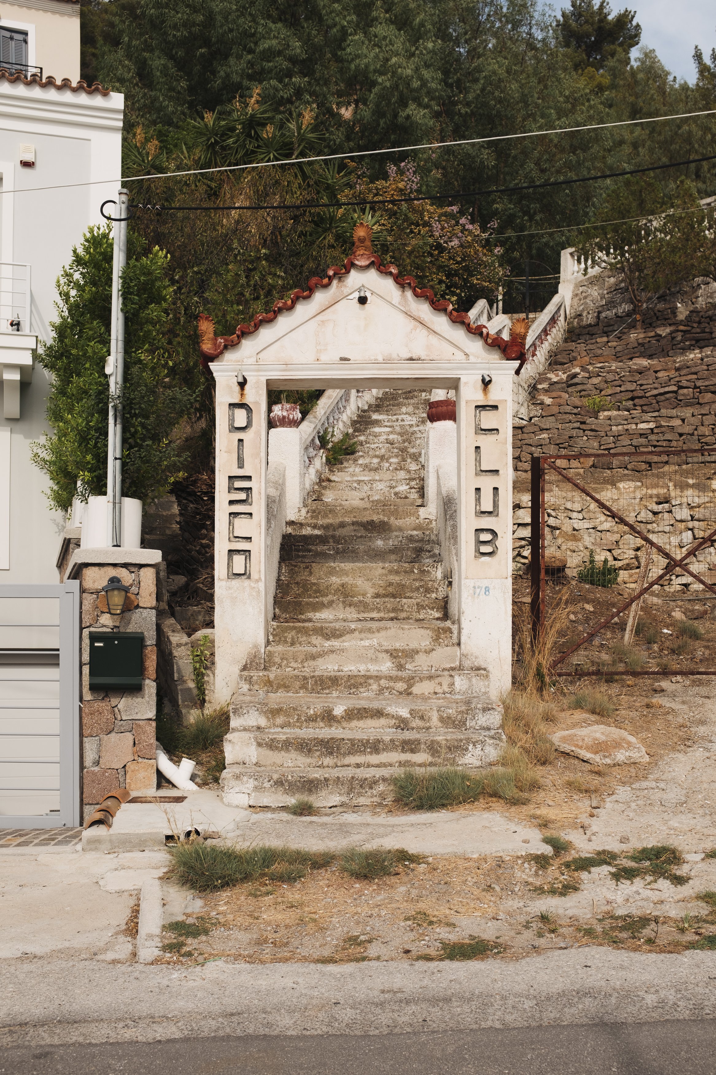 Concrete stairs leading up to a small tiled-roof archway with signage that reads 'Disco Club' on either side, situated on a sloped hill with rocks and greenery.