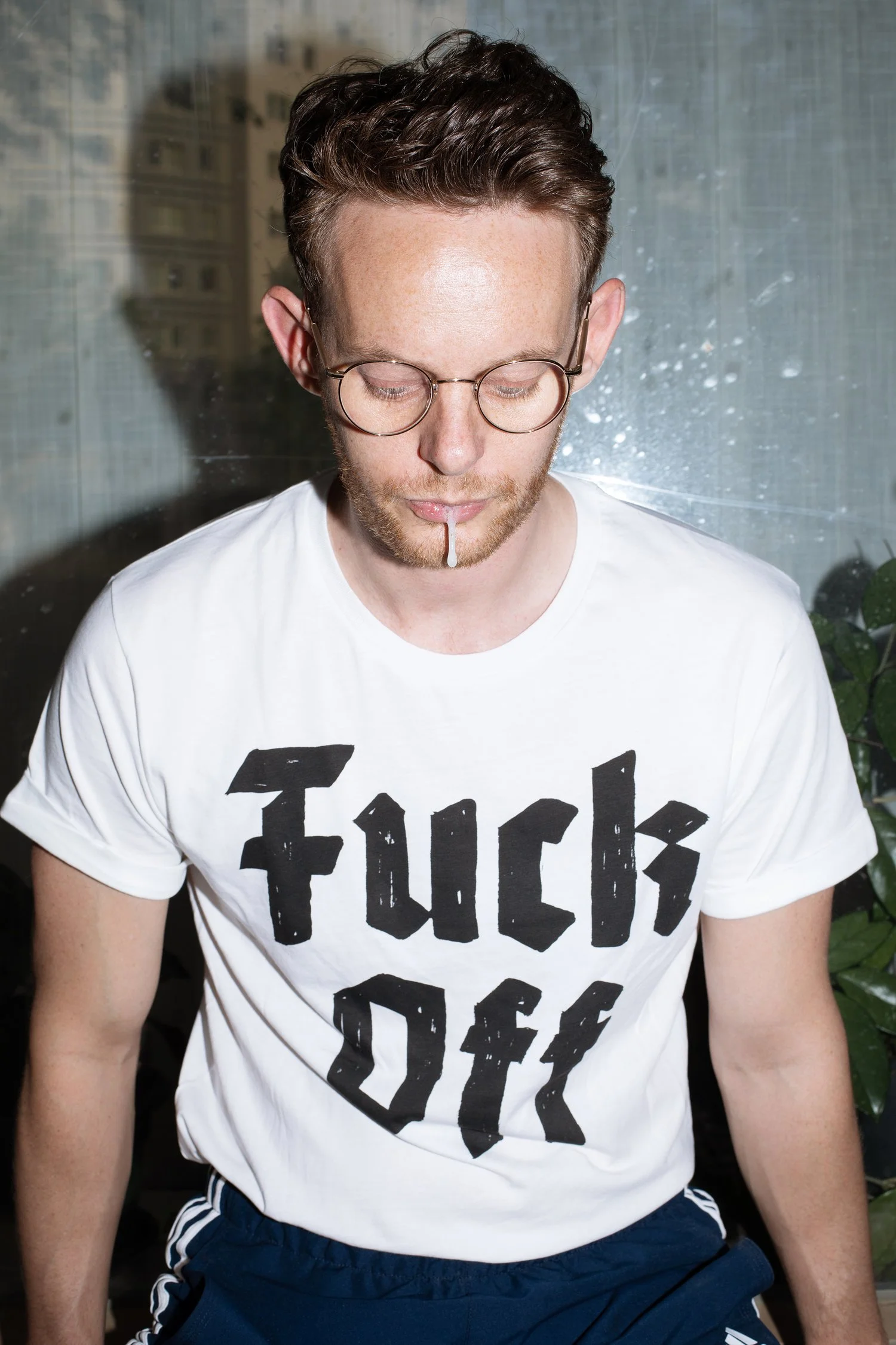 A young man with short brown hair, glasses, and a beard, wearing a white t-shirt with black text that says 'FUCK OFF,' is standing indoors with a blurred cityscape outside the window behind him. He is looking down with a cigarette hanging from his lips.