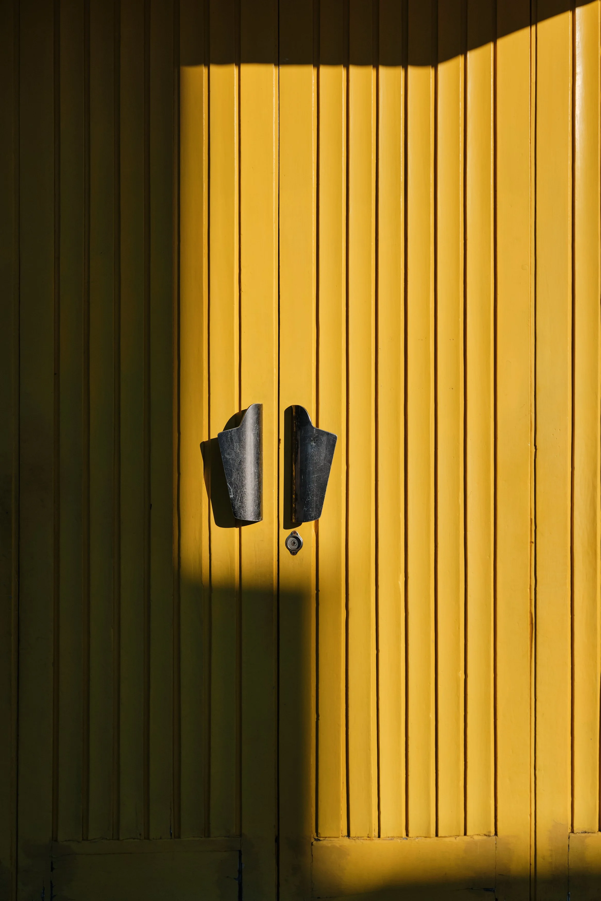 Yellow wooden double doors with black handles and a small lock underneath, partially lit by sunlight creating shadows.