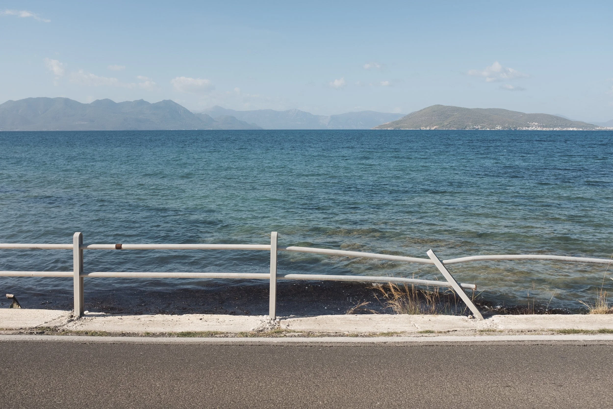View of a body of water with mountains in the distance and a metal guardrail along the shoreline in the foreground.