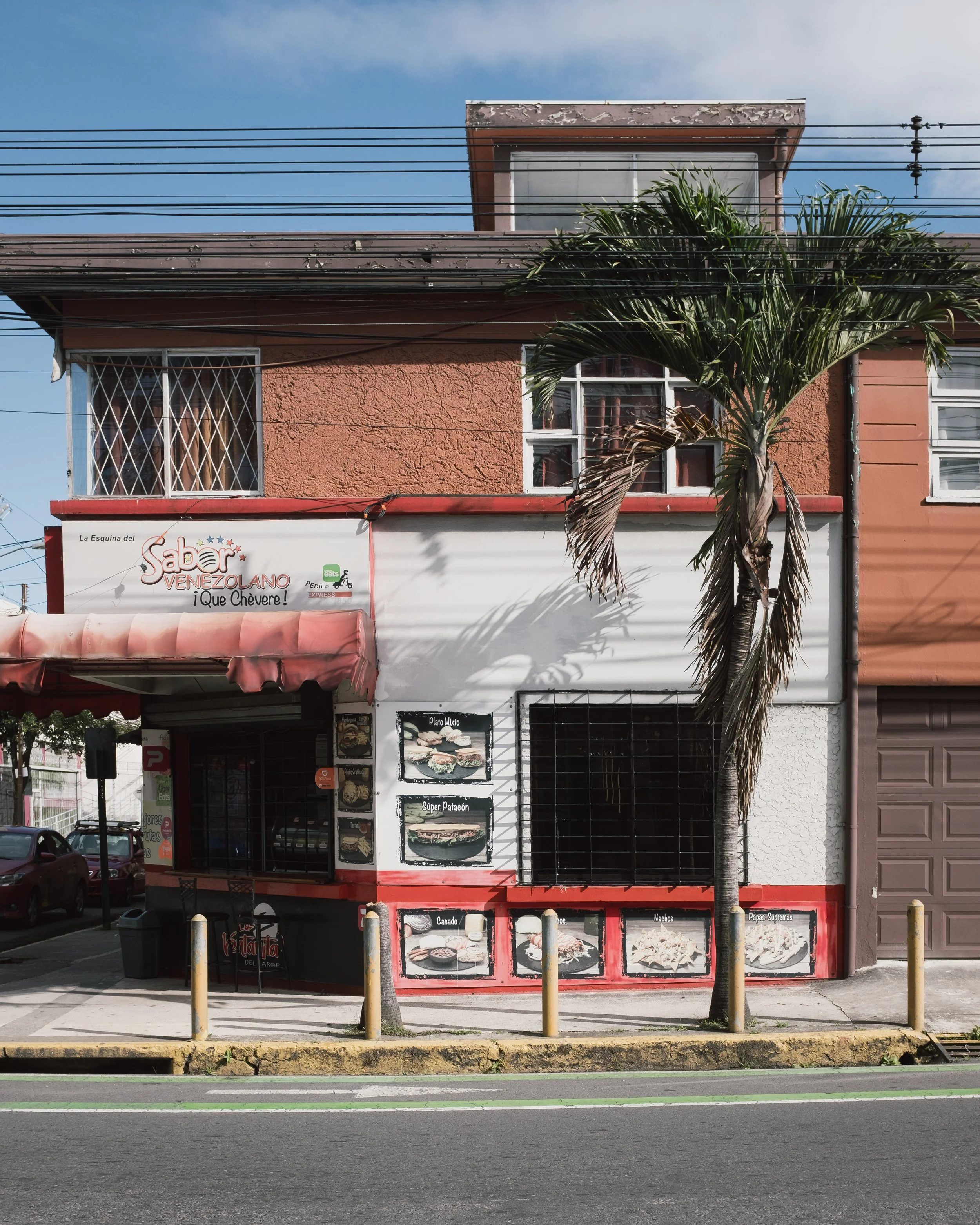 A small restaurant on a city street with signs in Spanish, displaying images of food items like sandwiches and snacks, with a palm tree in front, and a building with barred windows behind.