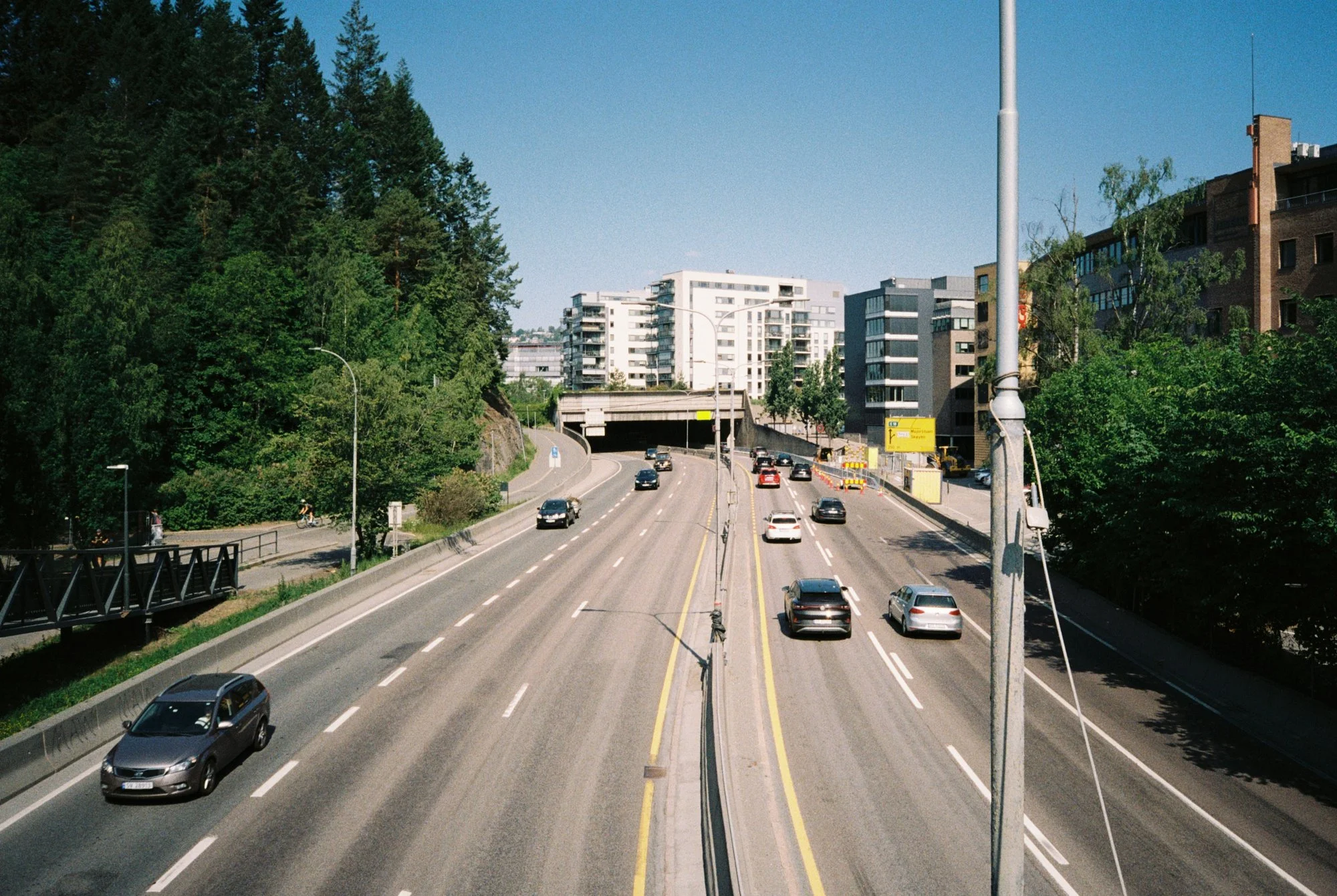 A multi-lane urban highway with several cars driving in both directions, bordered by tall green trees on the left and modern buildings on the right, under a clear blue sky.