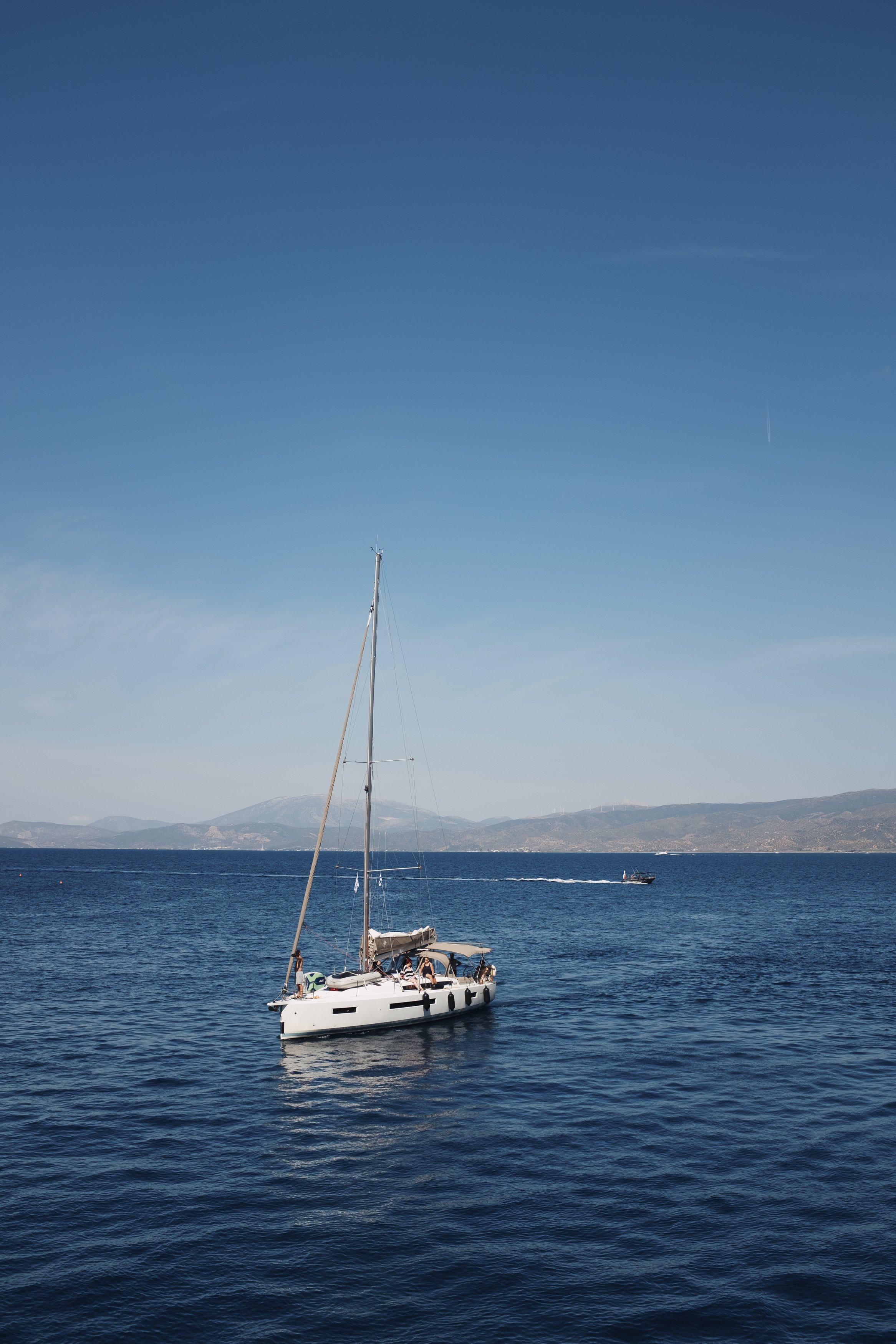 A sailboat with people on board floating on the ocean, with distant mountains and a clear blue sky in the background.