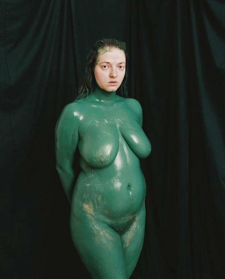 A woman covered in green body paint, standing against a black backdrop, with wet hair and a serious expression.