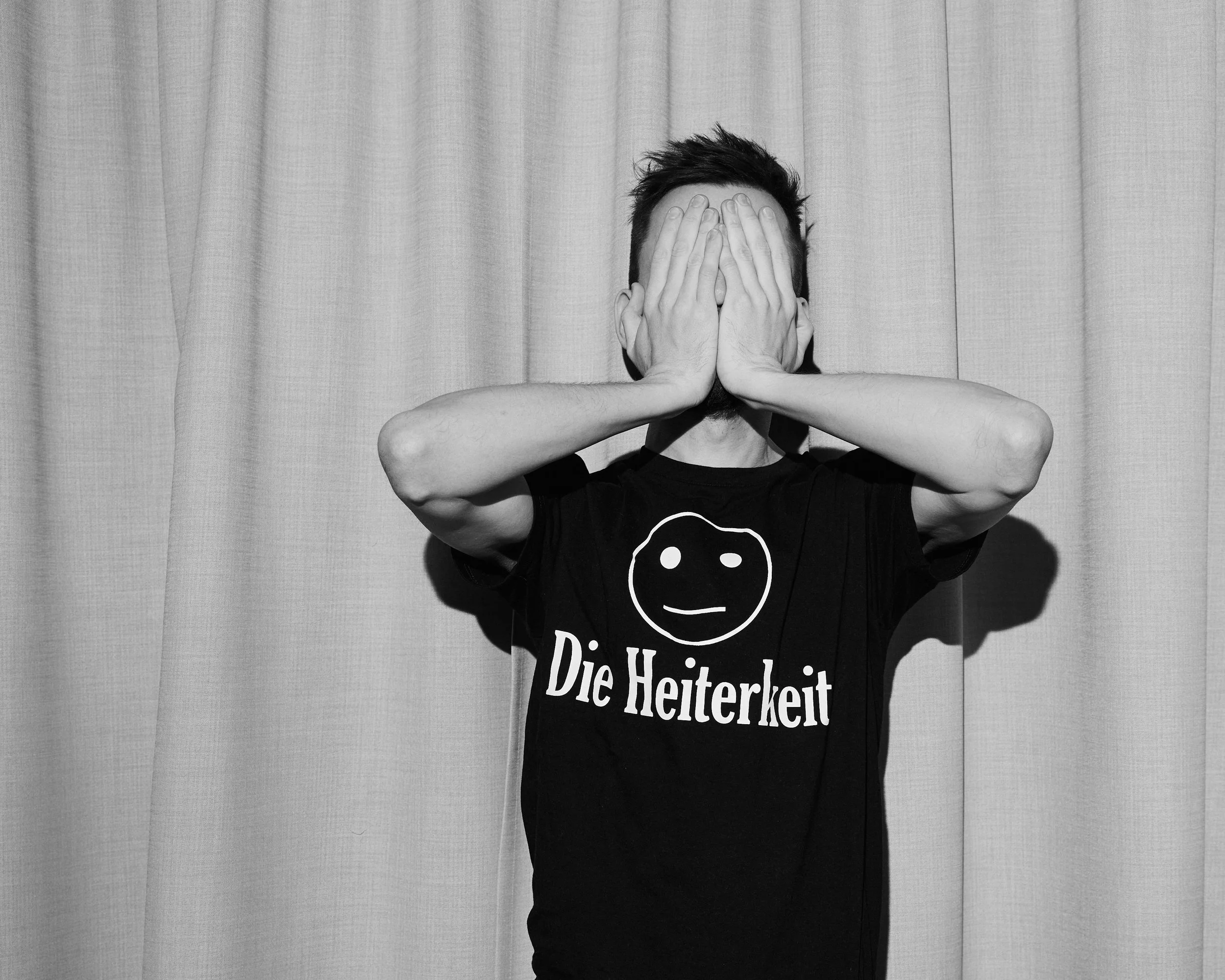 Black and white photo of a person standing in front of a curtain, covering their face with both hands. The person is wearing a T-shirt with a smiley face and the text "Die Heiterkeit."