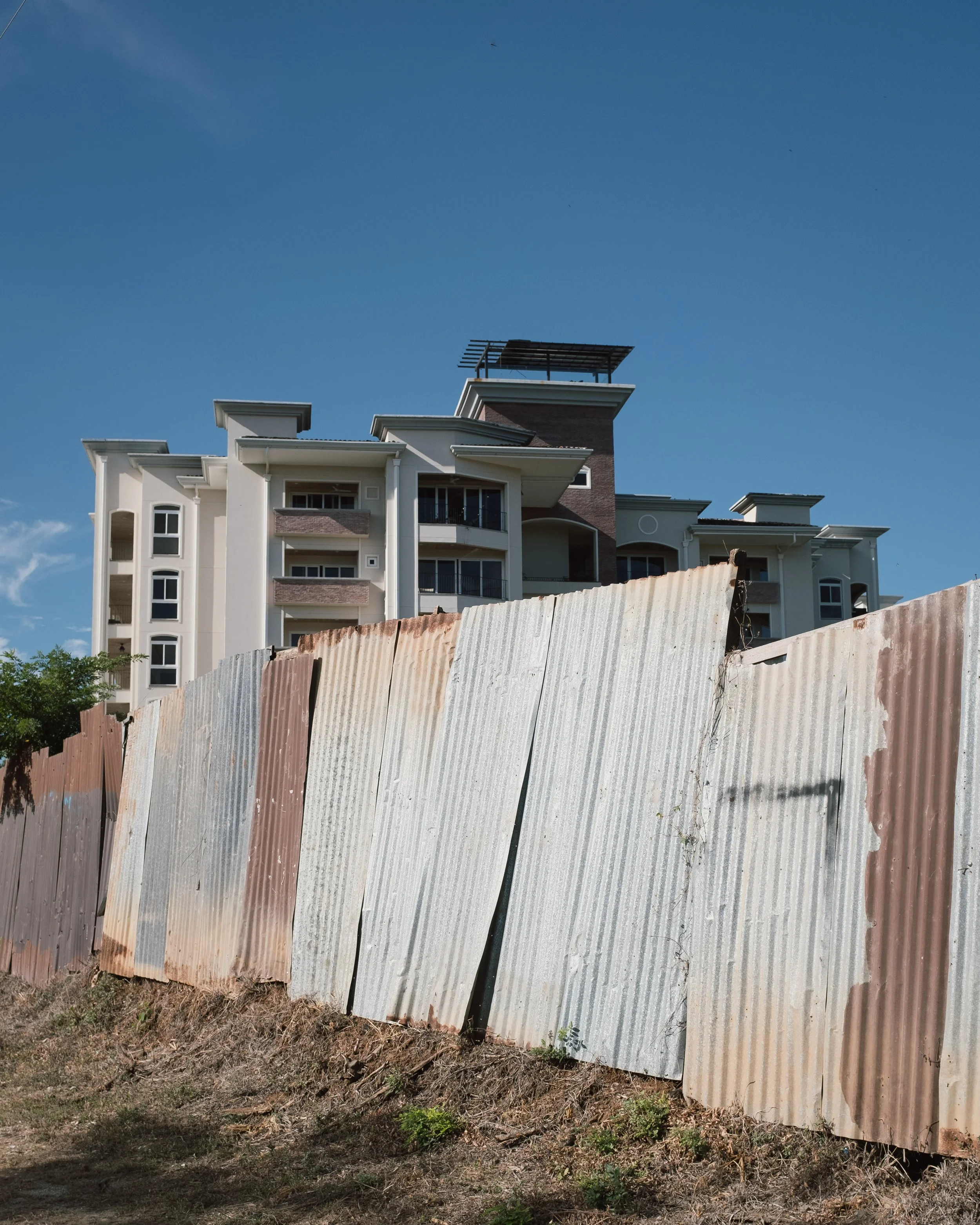 Rusted metal fence in foreground with a large modern residential building under a clear blue sky in the background.