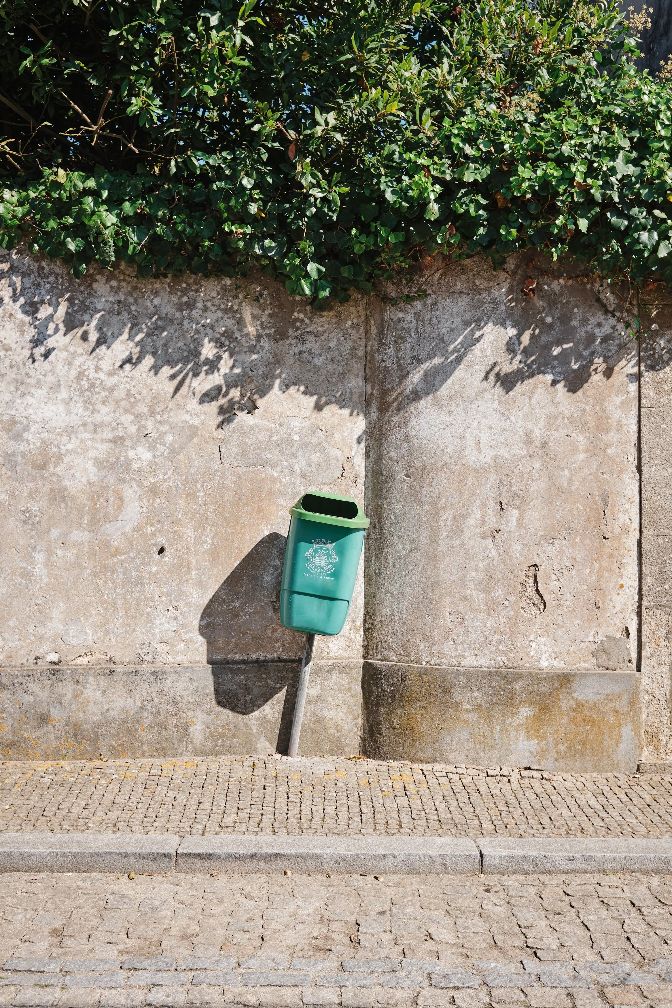 Green trash bin attached to a pole, mounted on a sidewalk with a stone wall and green bush overhead, casting a shadow.