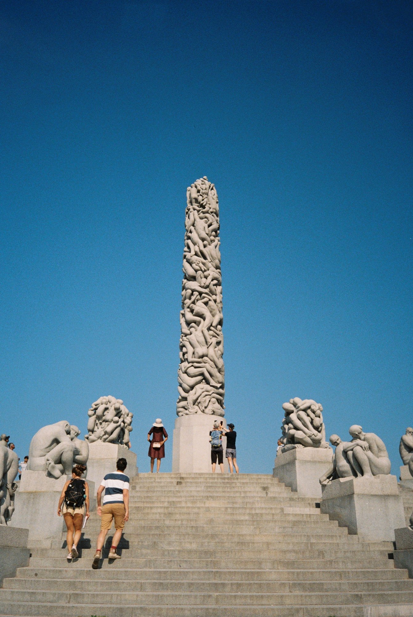 Group of tourists climbing stairs towards a tall, intricately carved stone monument with a clear blue sky in the background.