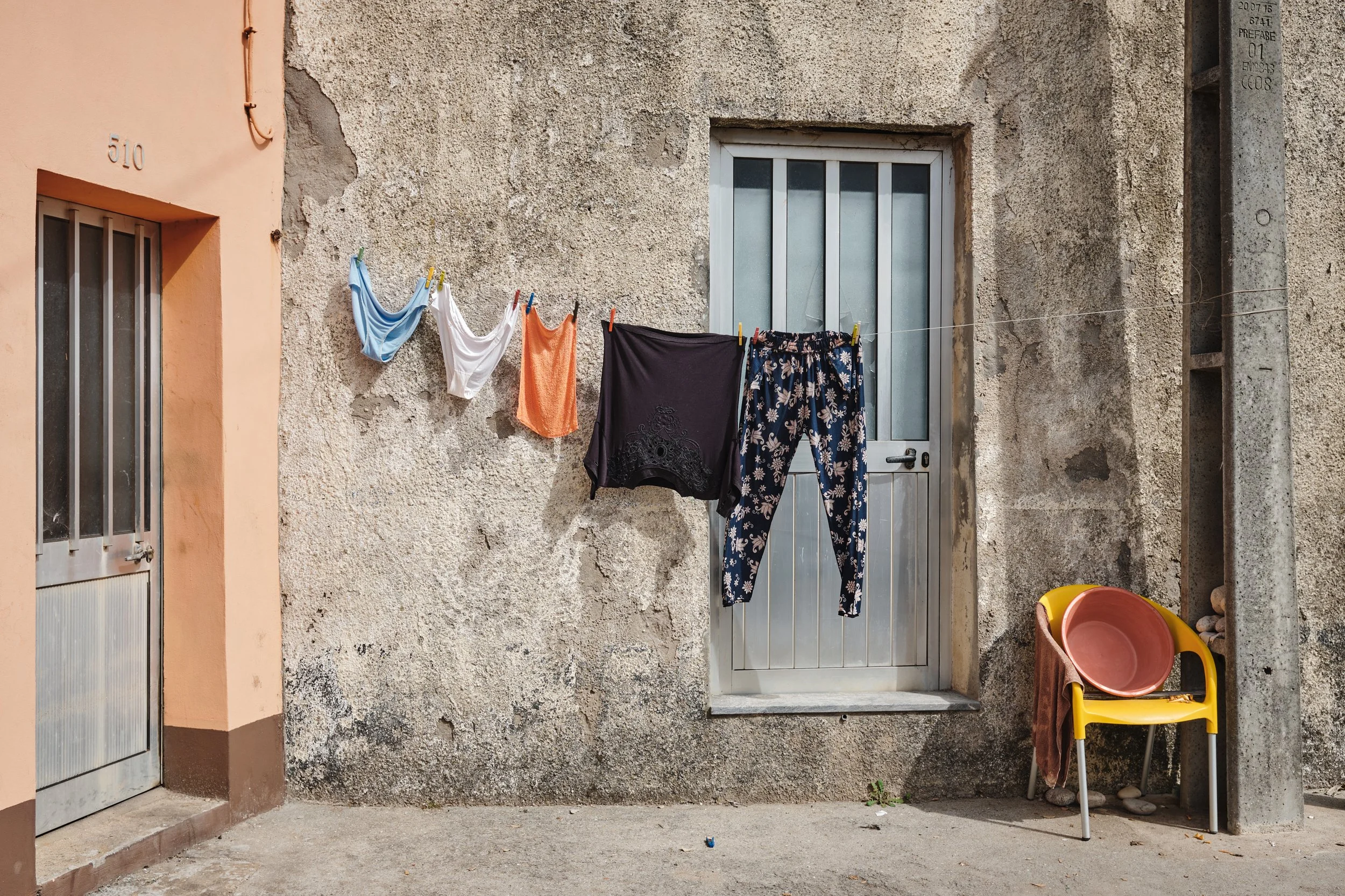 Clothes hanging on a clothesline outside a weathered stone wall with a door and window, and a yellow chair with a bowl and cloth beside a utility pole.