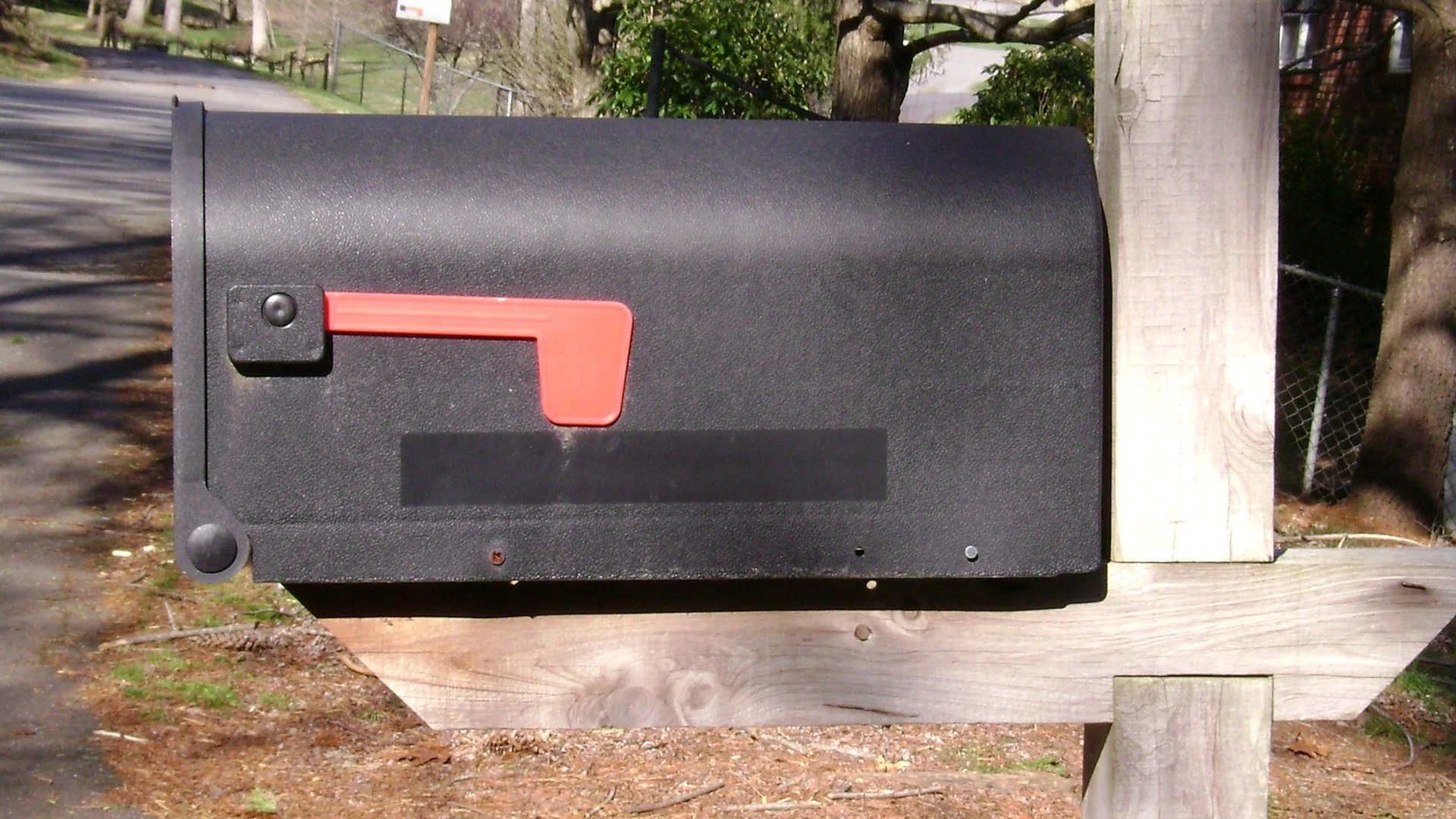 A black curbside mailbox with a red flag on a wooden post beside a quiet residential street
