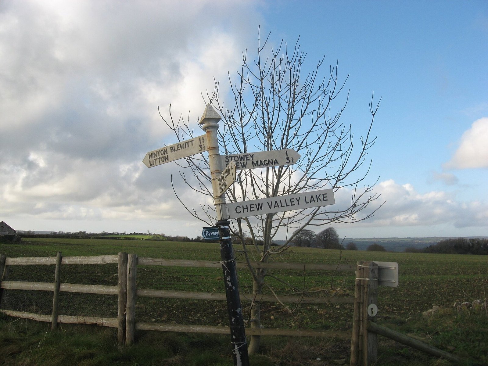Weathered wooden fingerpost at a rural crossroads with arms pointing in different directions under a cloudy sky