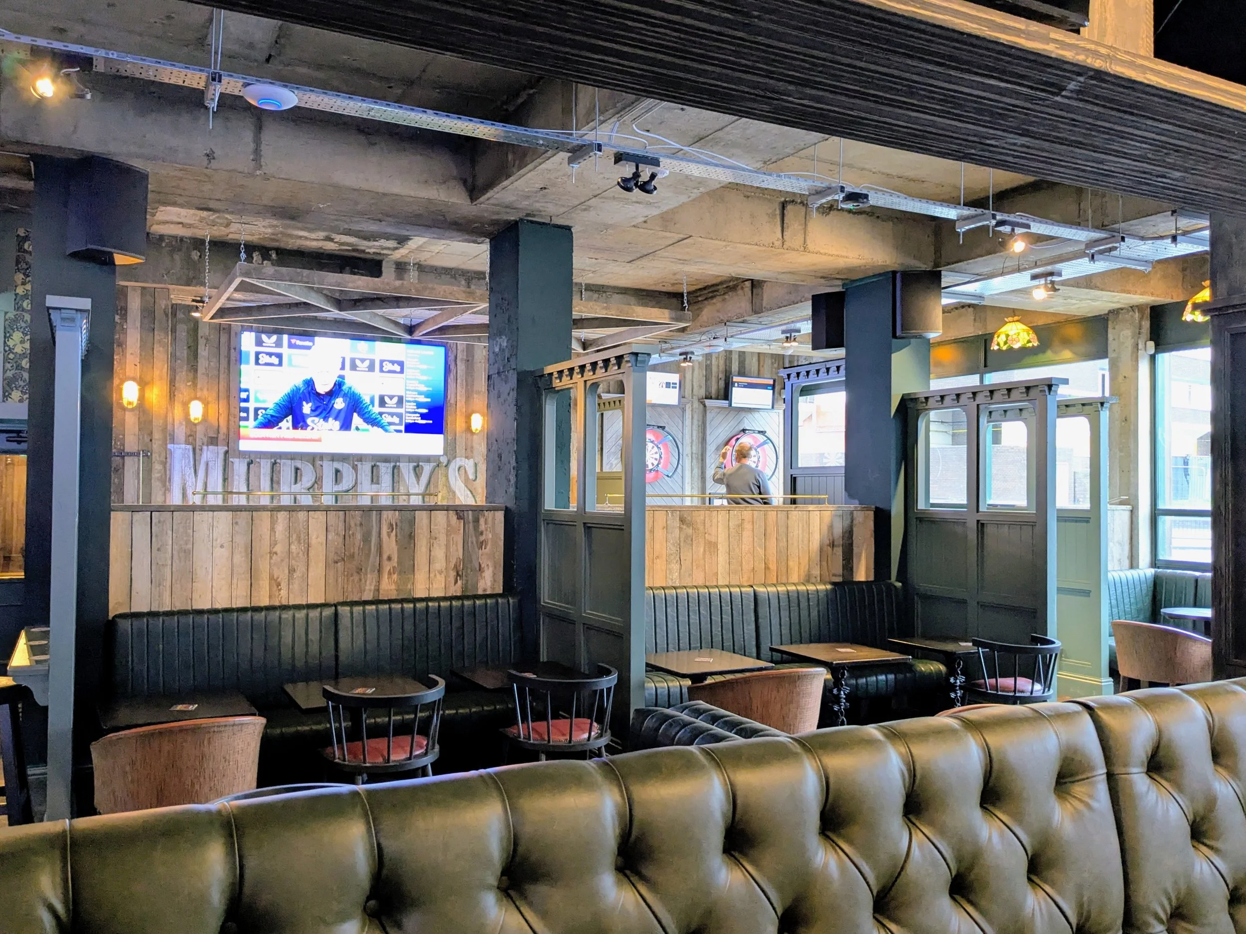 Interior of a bar or pub with leather seating, wooden booths, a large screen displaying a sports event, dartboards, and decorative lighting fixtures.